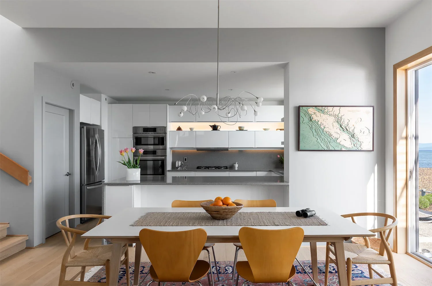 Modern kitchen with white cabinets, gray countertops, and a dining table with six wooden chairs. A bowl of oranges is on the table, and a vase of pink tulips is on the counter. There is a large window to the right showing an outdoor view of the ocean and sky.