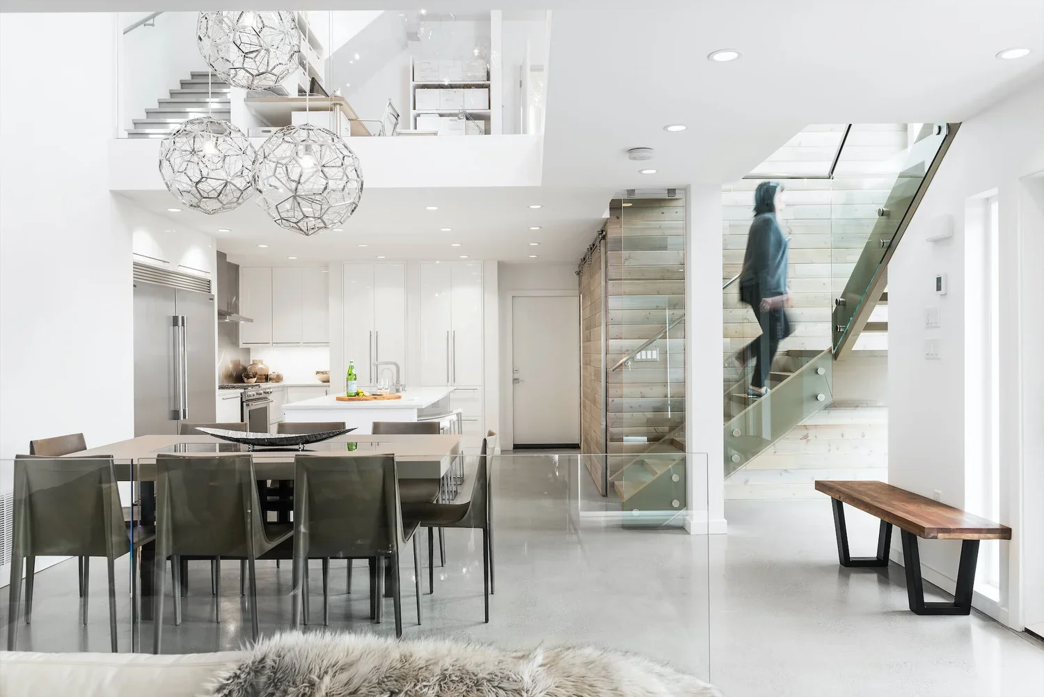 Modern, minimalist kitchen and dining area with a staircase. White cabinetry, stainless steel appliance, glass chandelier, and a person walking up the stairs.