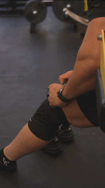A person sitting on gym floor with bent knees, wearing black shorts and black shoes, resting their arm on their thigh near weights in the background.