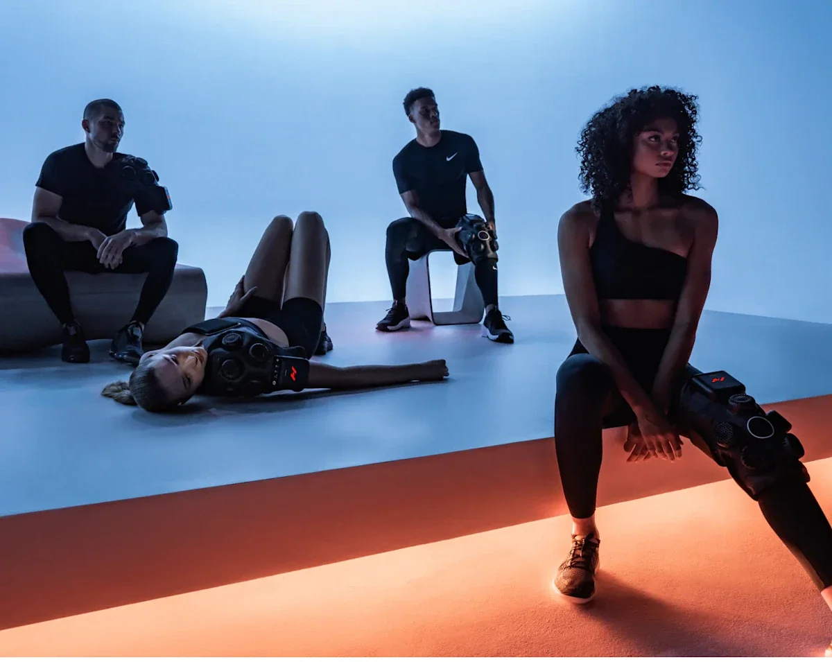 Four people in workout attire are in a fitness studio with colorful lighting, some sitting and one lying on the floor, all holding or wearing exercise equipment.