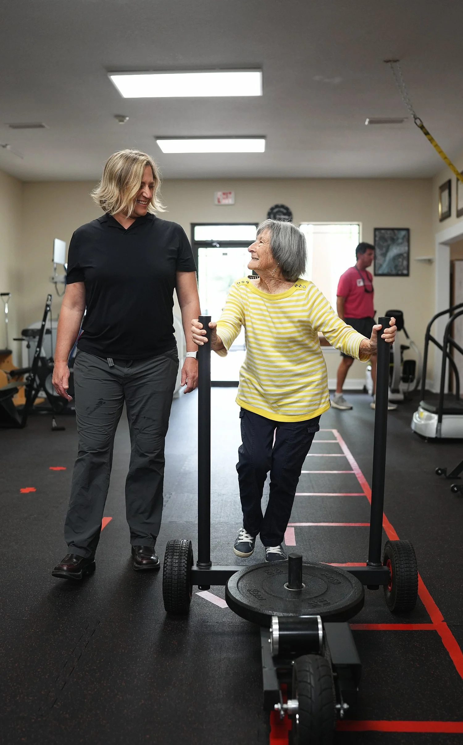 An elderly woman with gray hair and a yellow-striped shirt exercises with a sled push machine, assisted by a female trainer with shoulder-length blonde hair, in a gym while a man in a pink shirt supervises in the background.