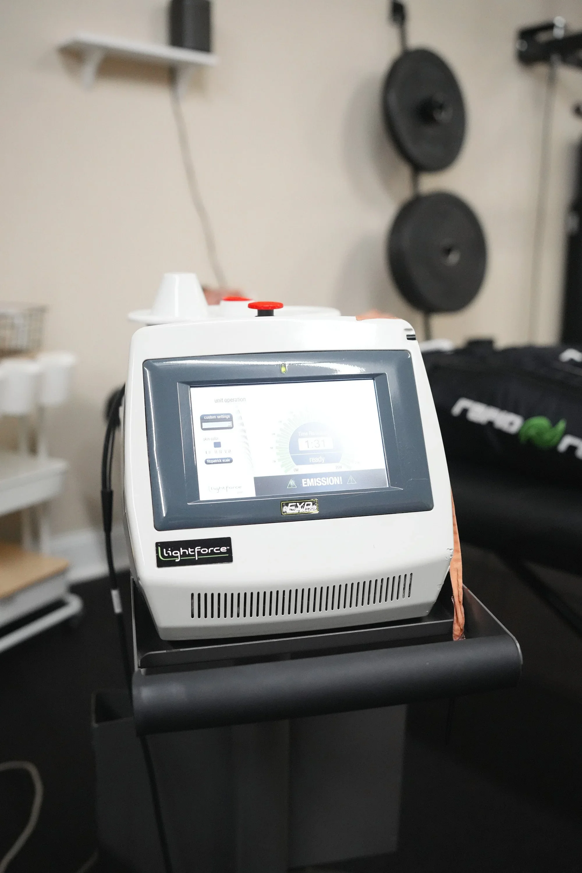 LightForce shockwave therapy machine with a digital display screen, mounted on a black stand in a gym or therapy room. Weights are visible in the background.