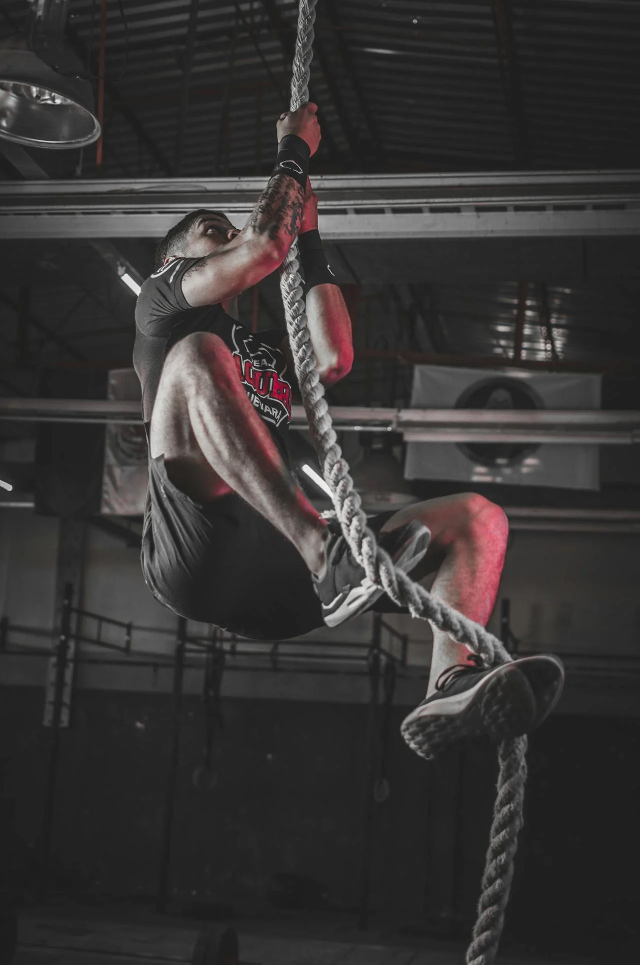 A man climbing a thick rope in a gym, wearing workout clothes and gloves, with gym equipment in the background.
