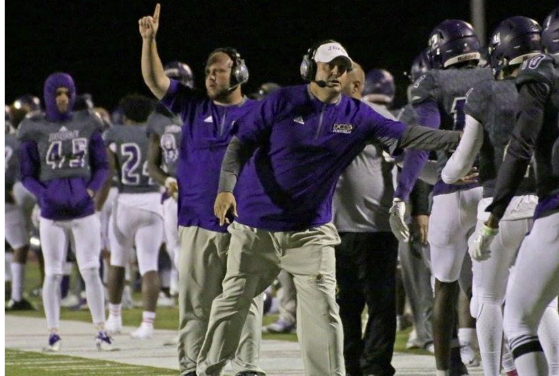 Football coach giving instructions on the sidelines during a game, with players in purple and white uniforms behind him.