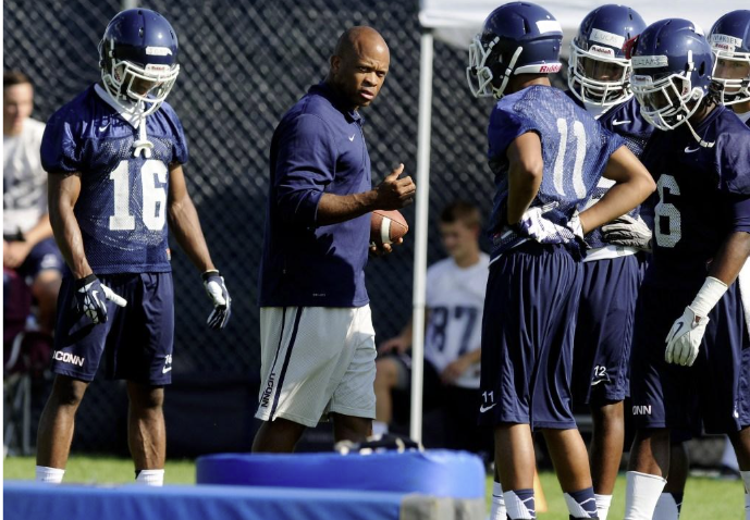 Football coach speaking to players during practice, players wearing navy blue uniforms and helmets, standing on field.