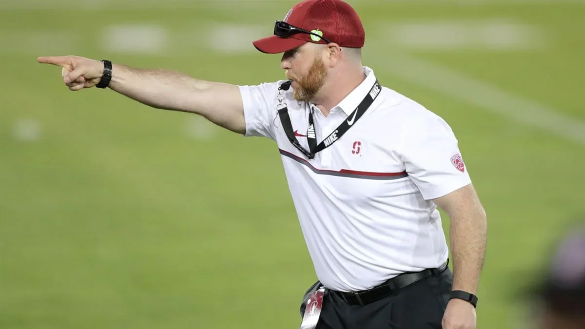 A football coach, wearing a white polo shirt, red cap, and sunglasses, is pointing and giving directions on the sideline of a football field.