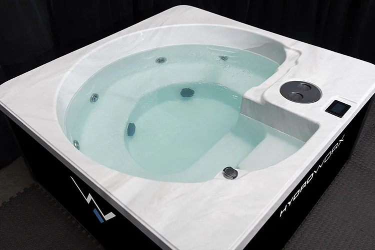 A white hot tub with built-in seats and jet nozzles filled with water, placed on a black textured floor.