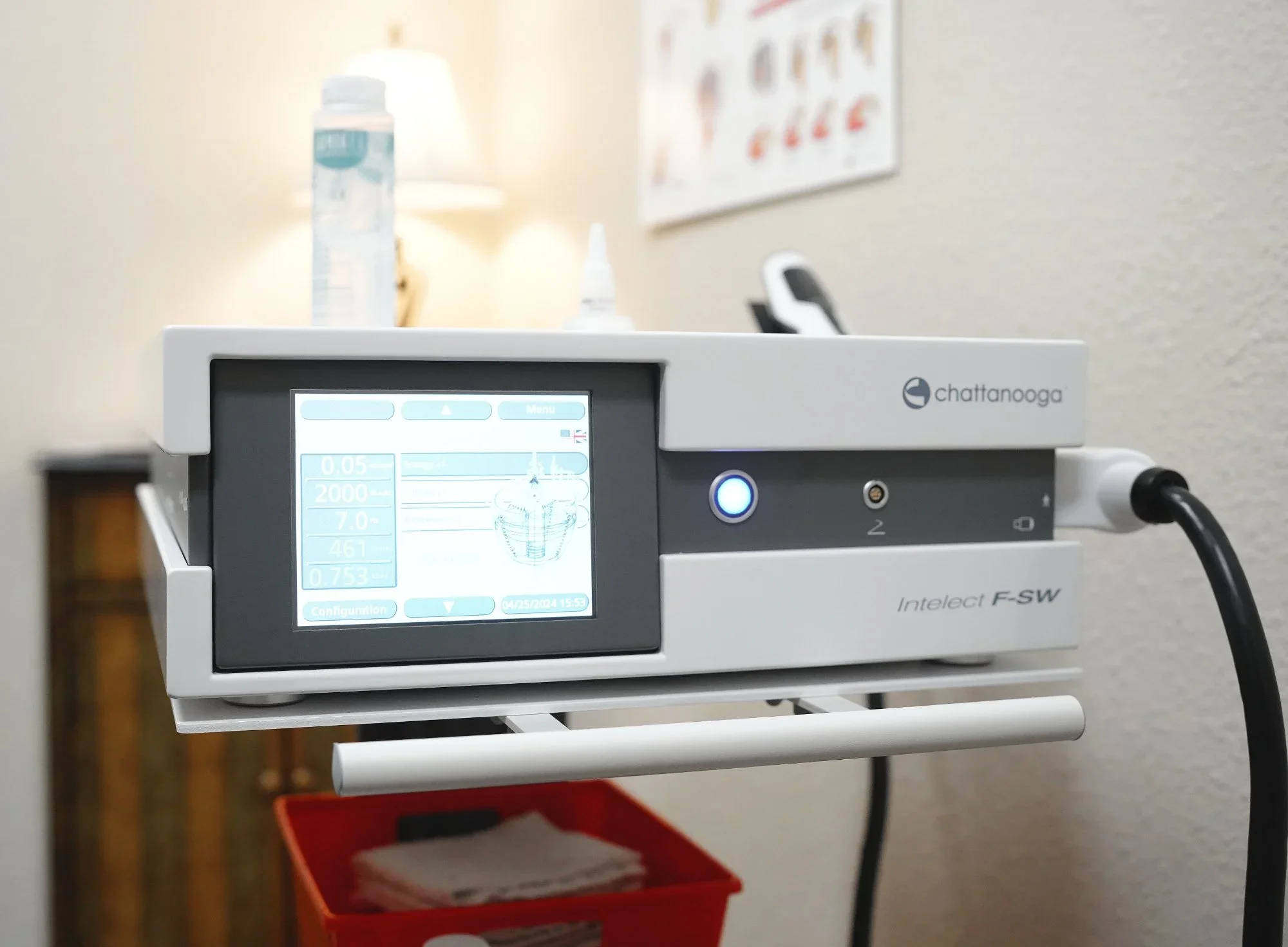 Medical device with a digital screen and a glowing blue button, connected to a power source, in a clinical setting.