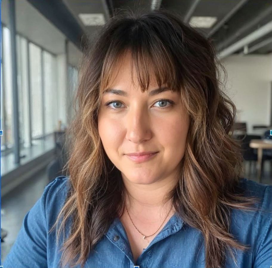A woman with wavy brown hair and blue eyes sitting indoors near large windows, wearing a blue shirt and a gold necklace.
