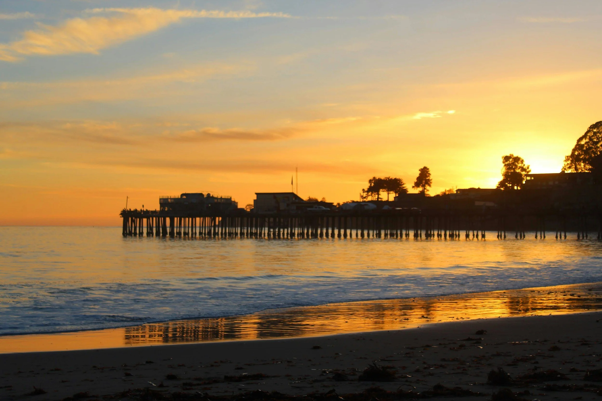 Sunset over a pier extending into the ocean, with silhouetted buildings and trees on the shoreline, reflecting warm orange and yellow hues on the water.