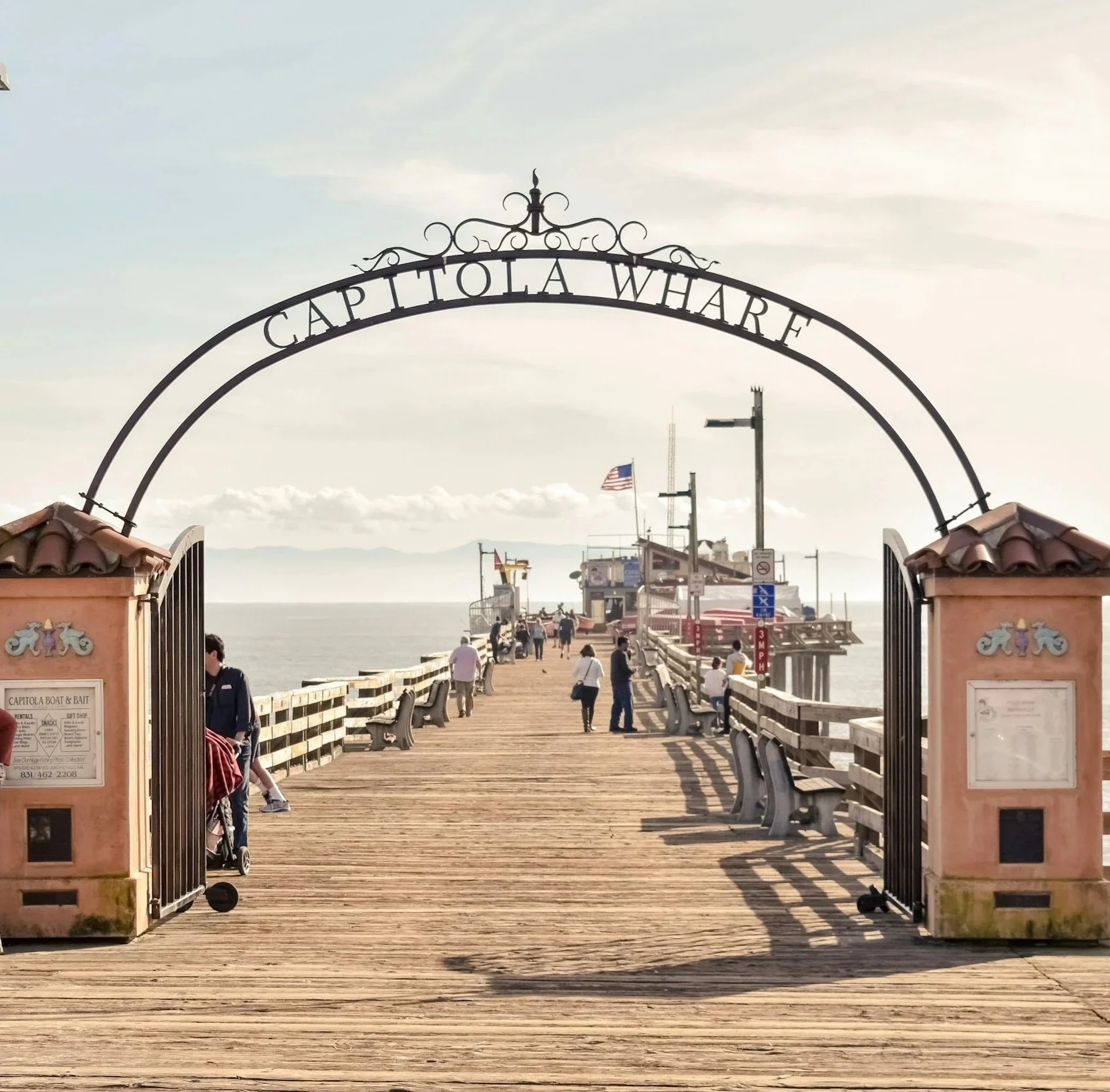 People walking on a wooden pier leading to a boat dock at Capo la White, with chairs and signage on the sides, and the ocean in the background.