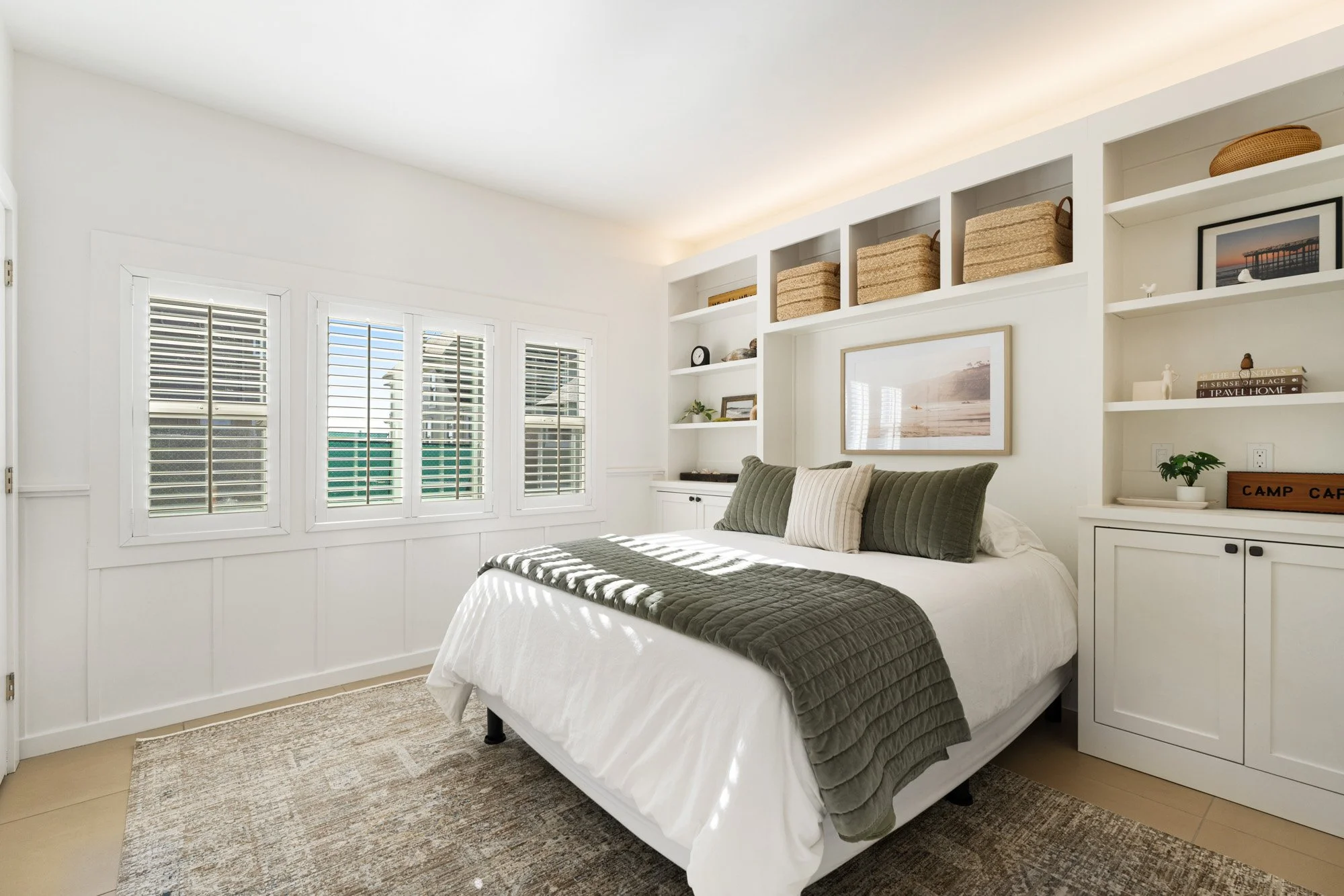 Bright bedroom with white walls, covered with white and green bedding, built-in shelves with baskets and decorative items, and a window with white plantation shutters.