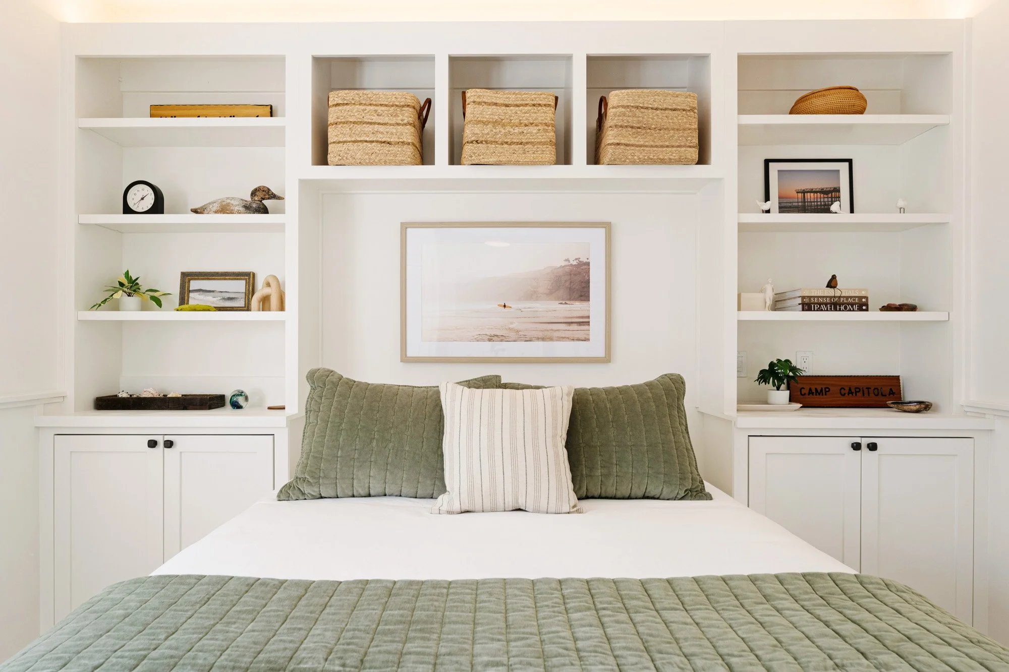 A cozy bedroom with a bed featuring green quilted pillows and a white-striped pillow. Behind the bed is a framed landscape photograph. White built-in shelves above the bed hold decorative items, books, and baskets, with some shelves in square compart