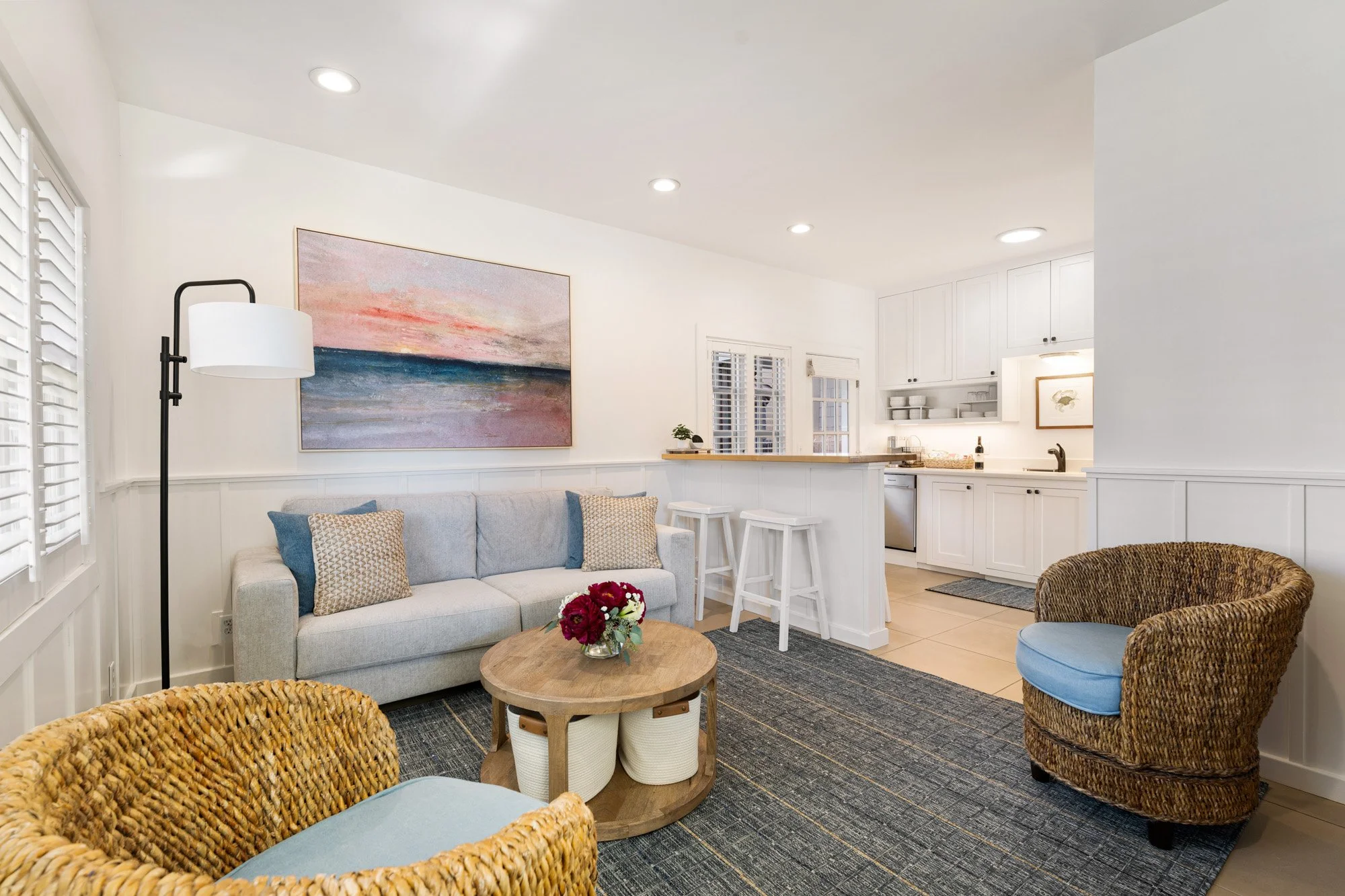 Living room with a white sofa, wicker chairs with blue cushions, a round wooden coffee table with a flower bouquet, and a patterned area rug. A floor lamp stands beside the sofa, and a large abstract painting hangs above it. The room opens to a kitch