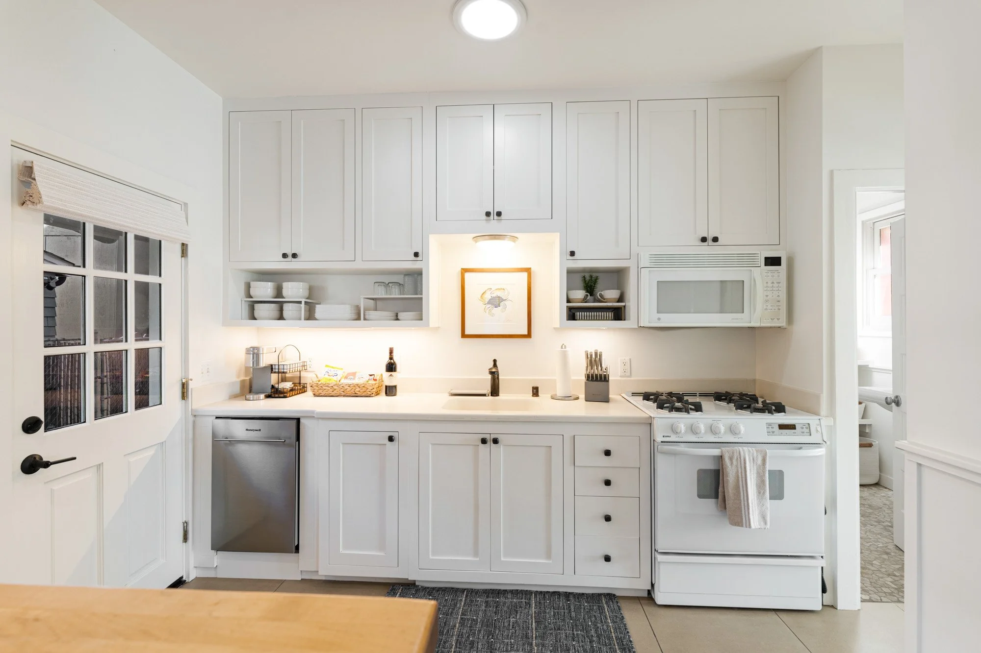 White kitchen with upper and lower cabinets, a built-in microwave, a stove, a small refrigerator, open shelves with dishes, a framed picture, and a door with glass panels.