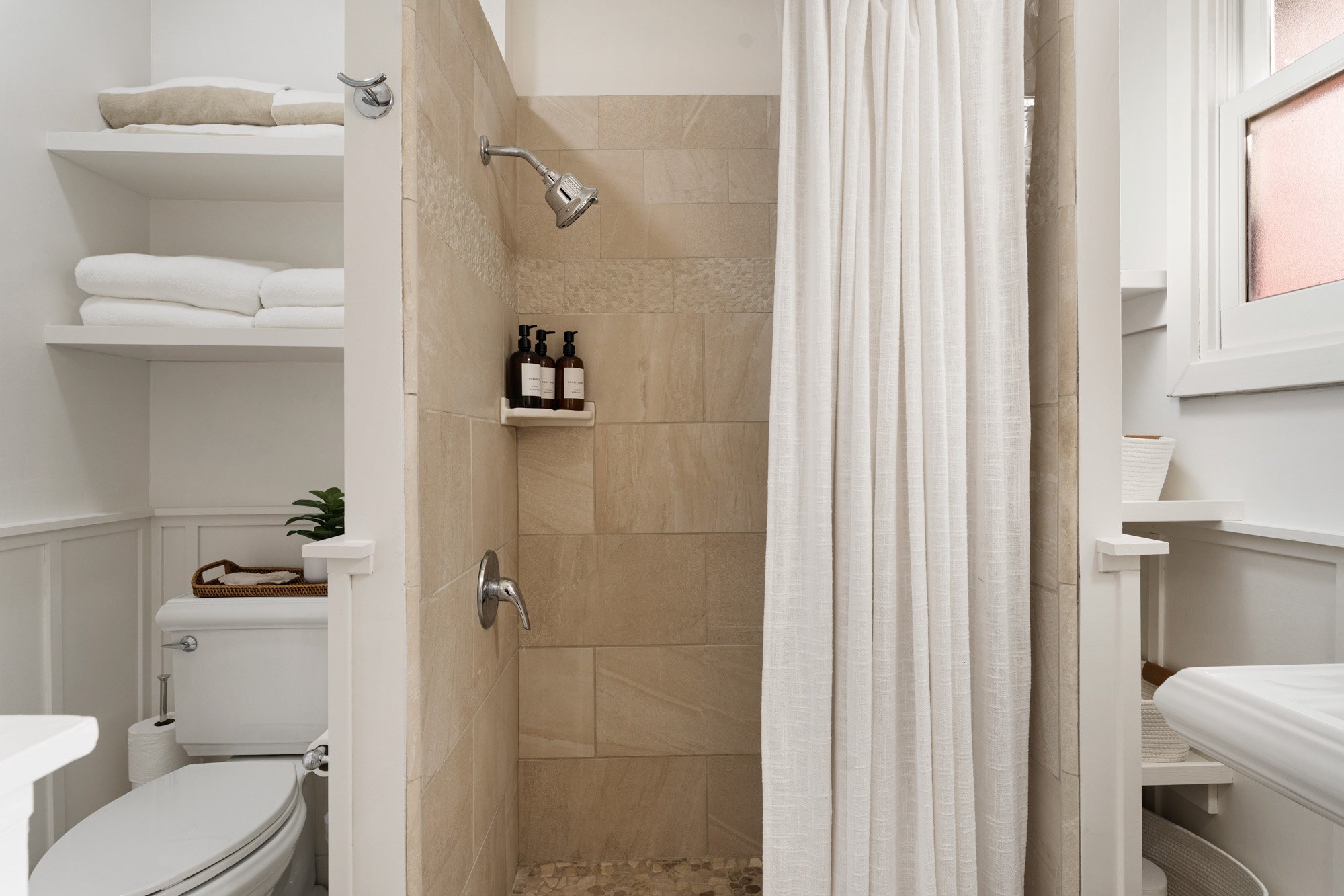 Small bathroom with a shower area enclosed by a white curtain, beige tiles, and shelves with toiletries, a toilet with a basket, towels, and a window.
