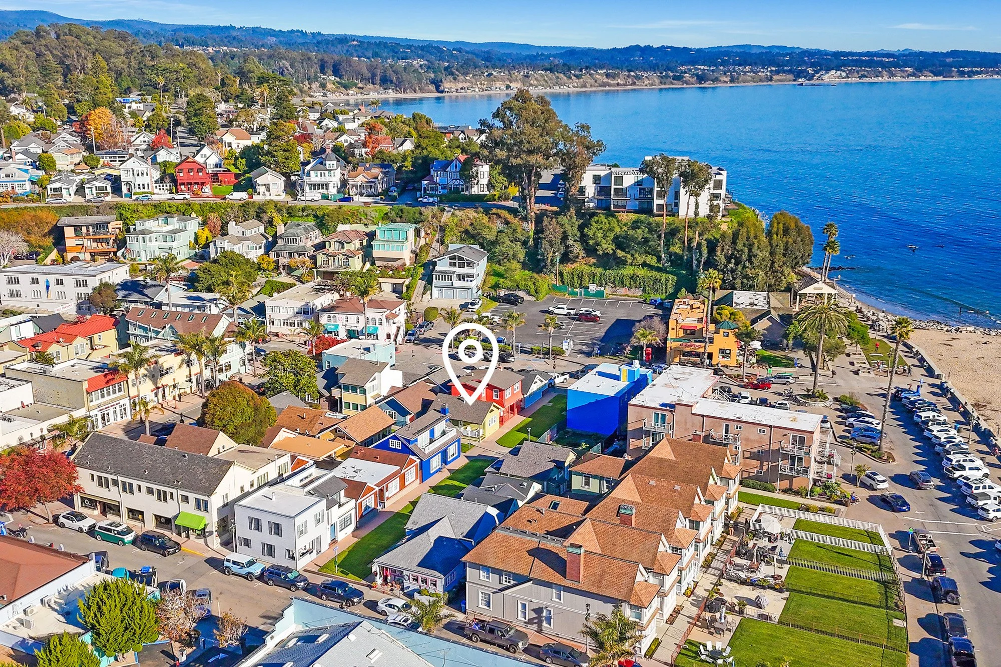 Aerial view of a coastal neighborhood with colorful houses, a parking lot, and a beach along the ocean with clear blue water.