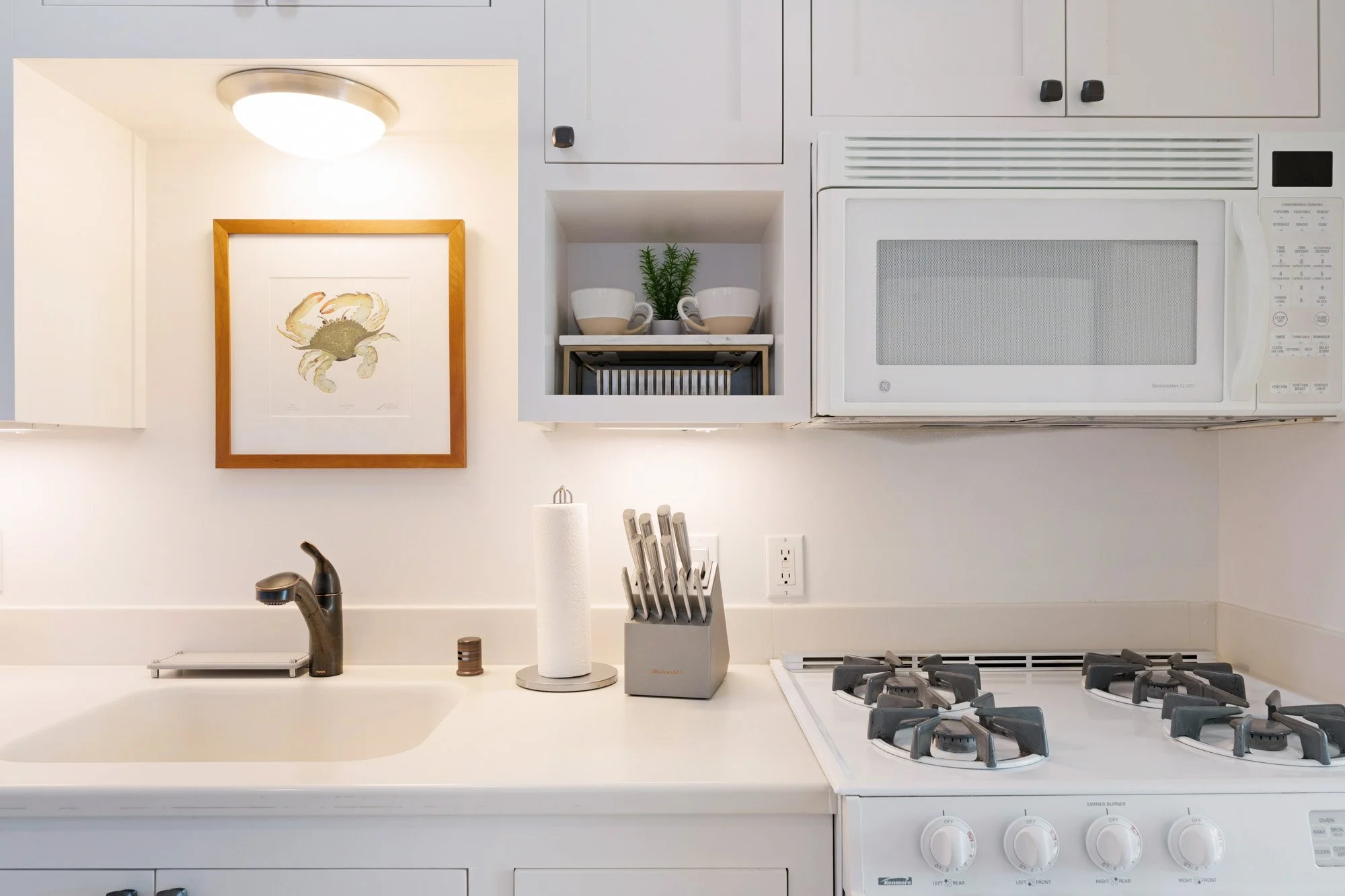 White kitchen with sink, gold crab print in wooden frame, paper towel roll, knife block, stove with knobs, microwave, cabinets, and small potted plant.
