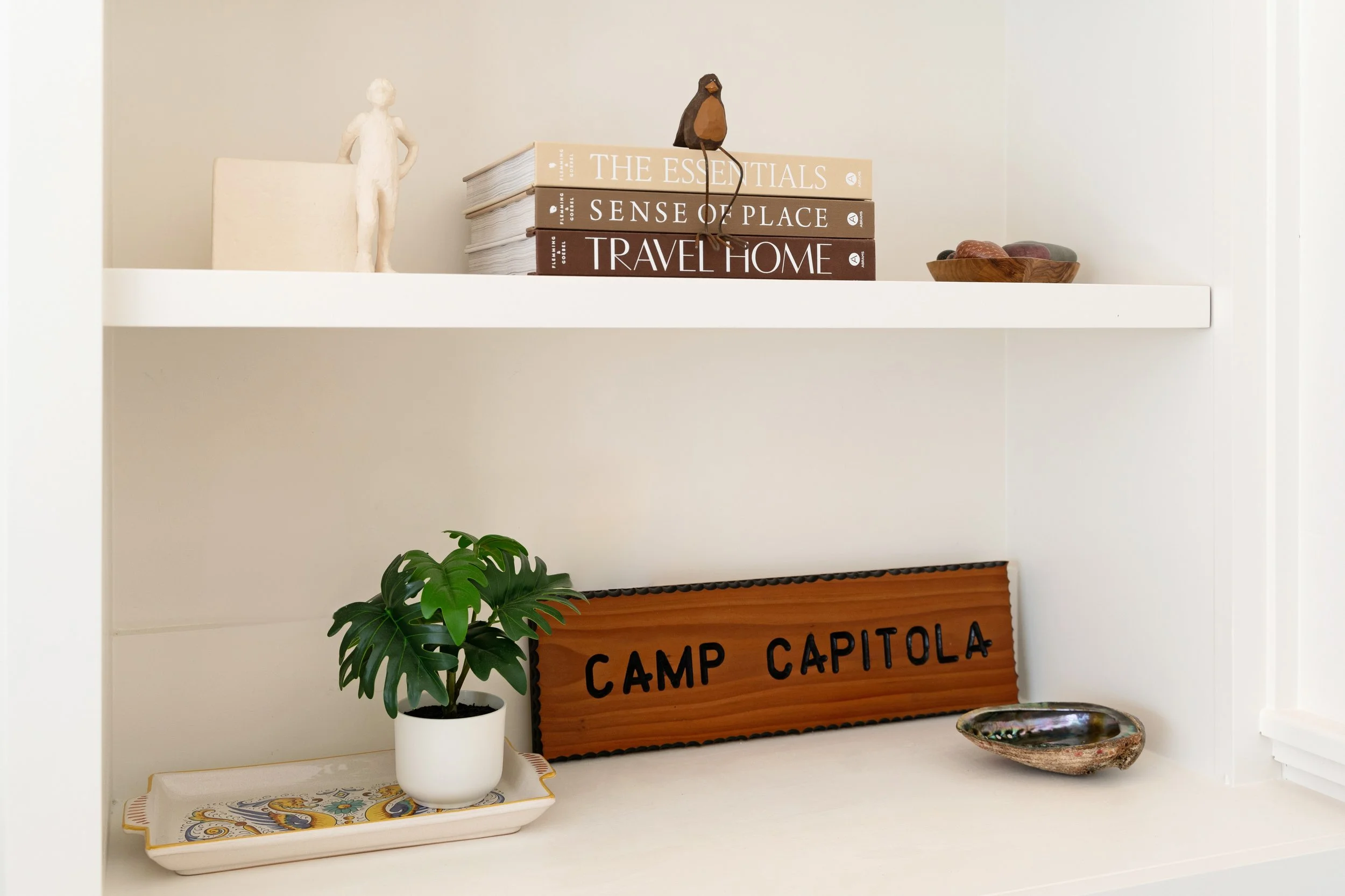 Decorative white shelf with books, a small sculpture of a person, a bird figurine, a wooden bowl, a potted plant on a tray, a wooden sign reading 'CAMP CAPITOLA', and a decorative ceramic bowl.