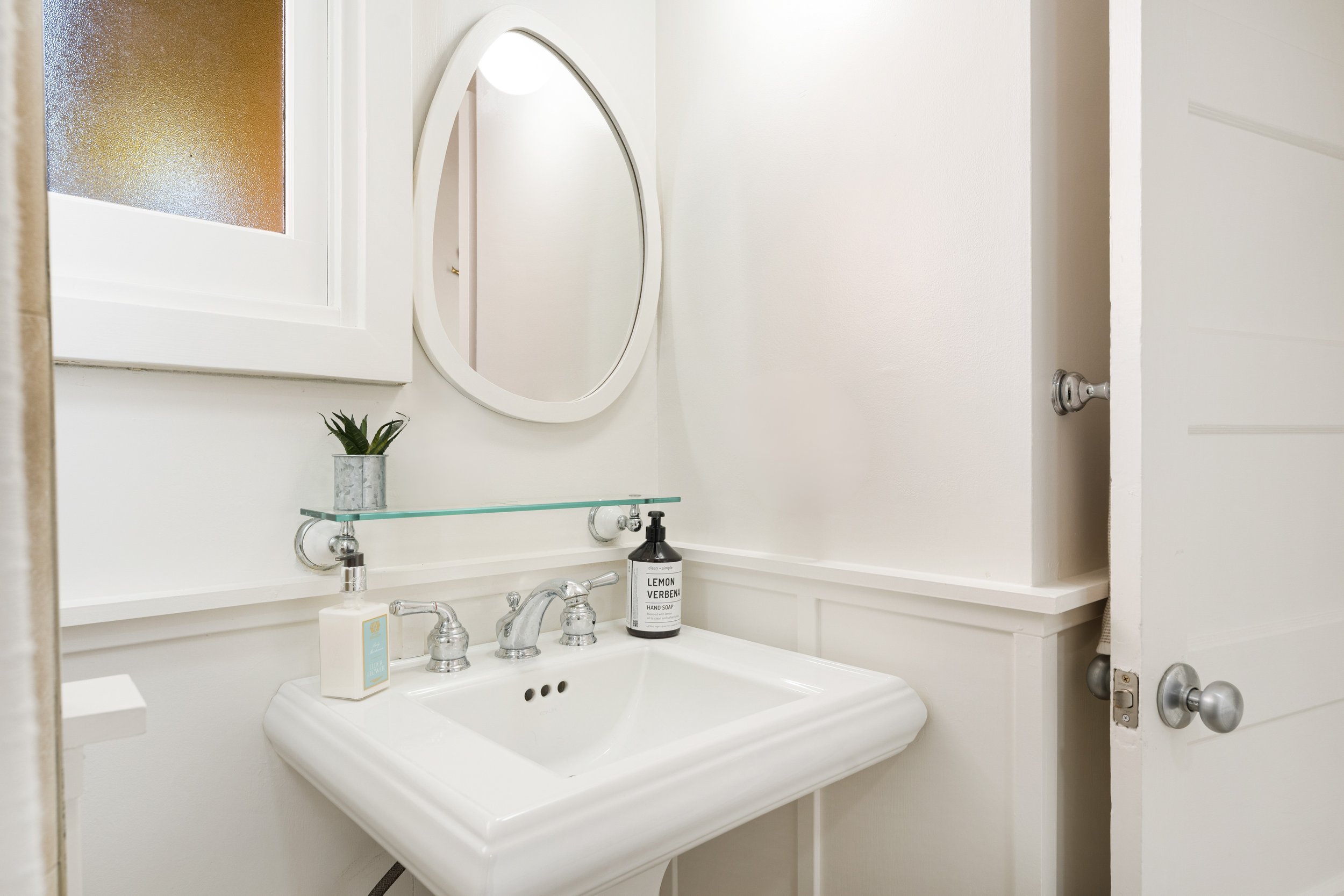 A small bathroom with a white sink, a round mirror, a potted plant, hand soap, and a glass shelf above the sink. The door is slightly open, showing part of a white wall and door handle.