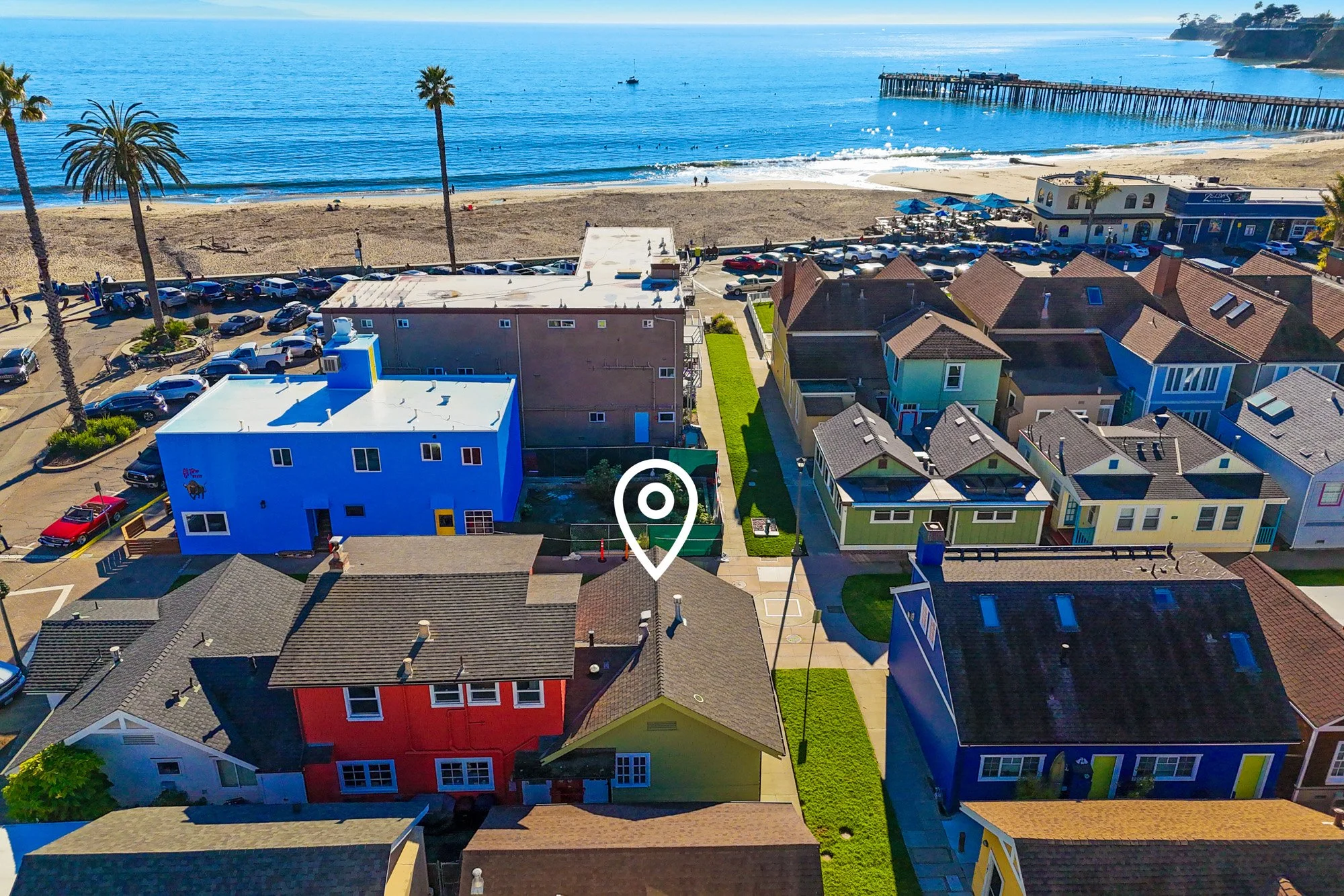 Aerial view of a coastal neighborhood with colorful houses, a parking lot, and a beach with palm trees and a pier extending into the ocean.