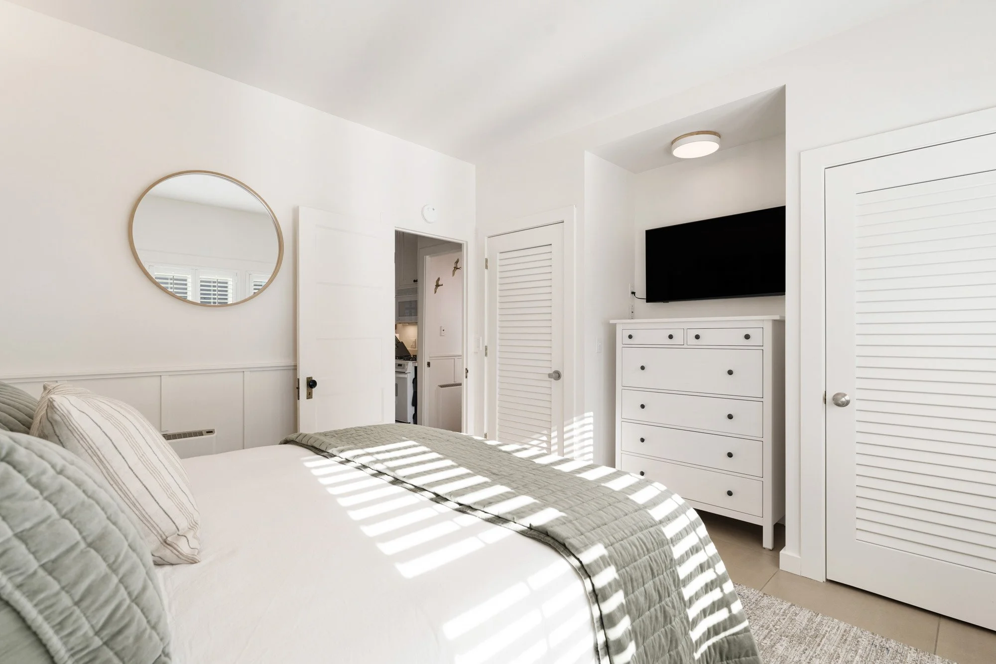 Bright bedroom with white walls, a bed with beige and gray bedding, a mirror on the wall, a dresser with a TV, and sunlight creating striped shadows on the bed.