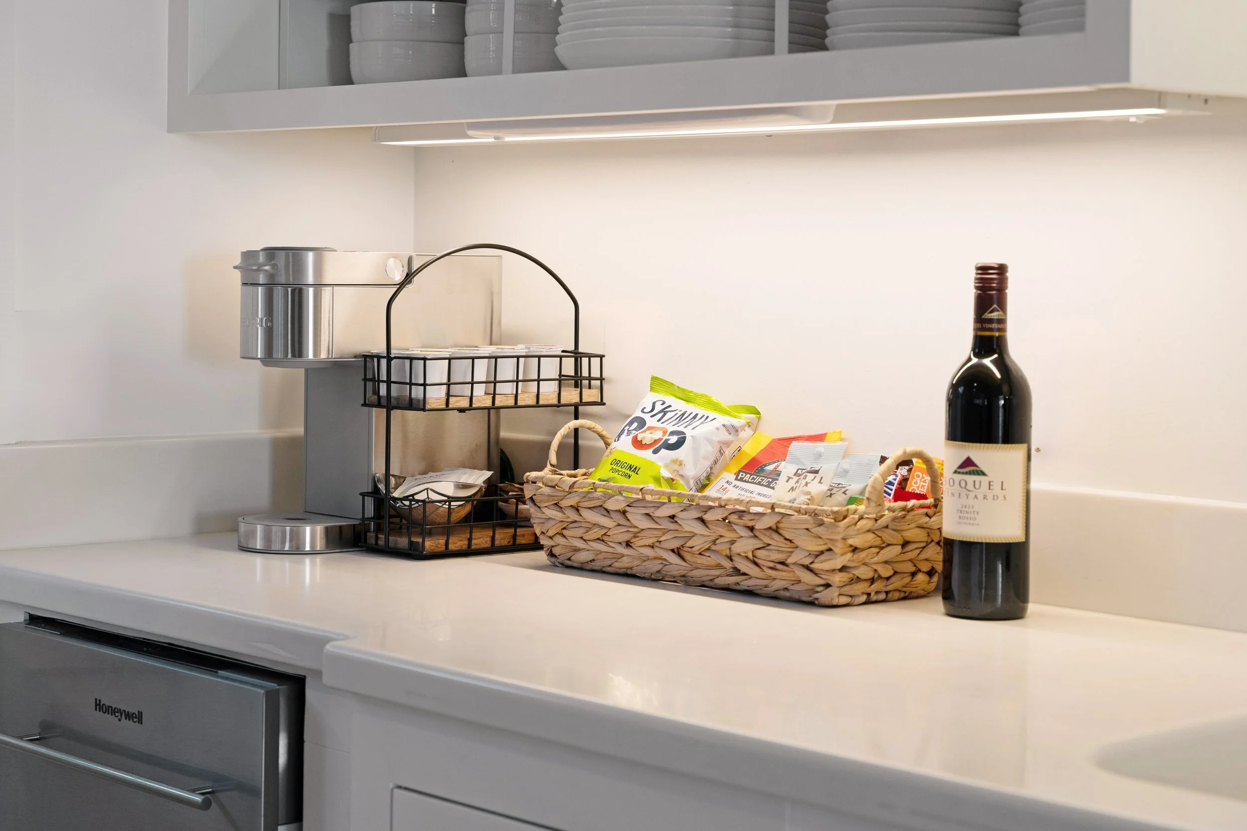 Kitchen countertop with a bottle of red wine, a woven basket filled with snack wrappers, a black wire dish rack, and a coffee machine in the background.
