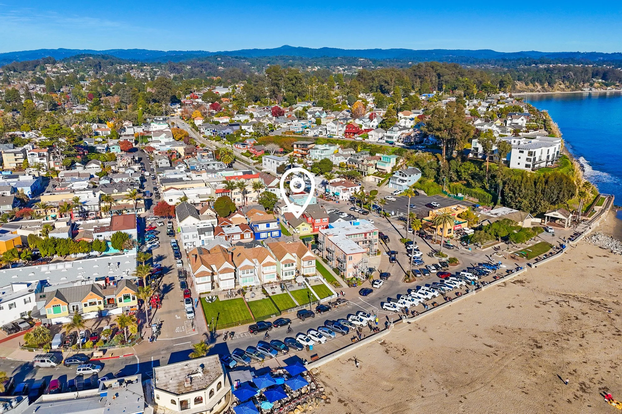 Aerial view of a coastal town with colorful houses, parking lots, and a beach, next to a large body of water with hills in the background.