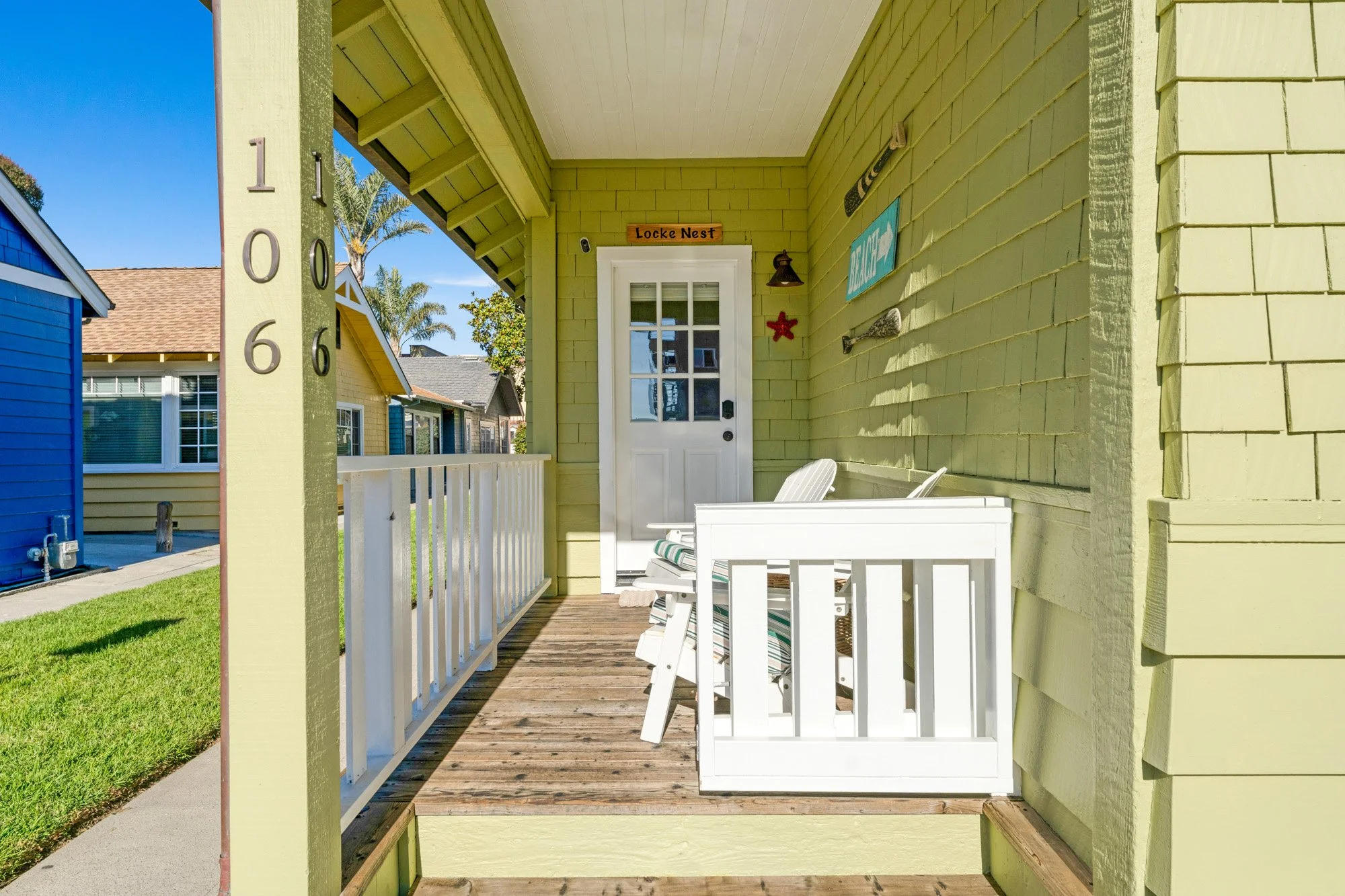 Front porch of a yellow house with a white door, white railing, Adirondack chairs, and nautical decorations.