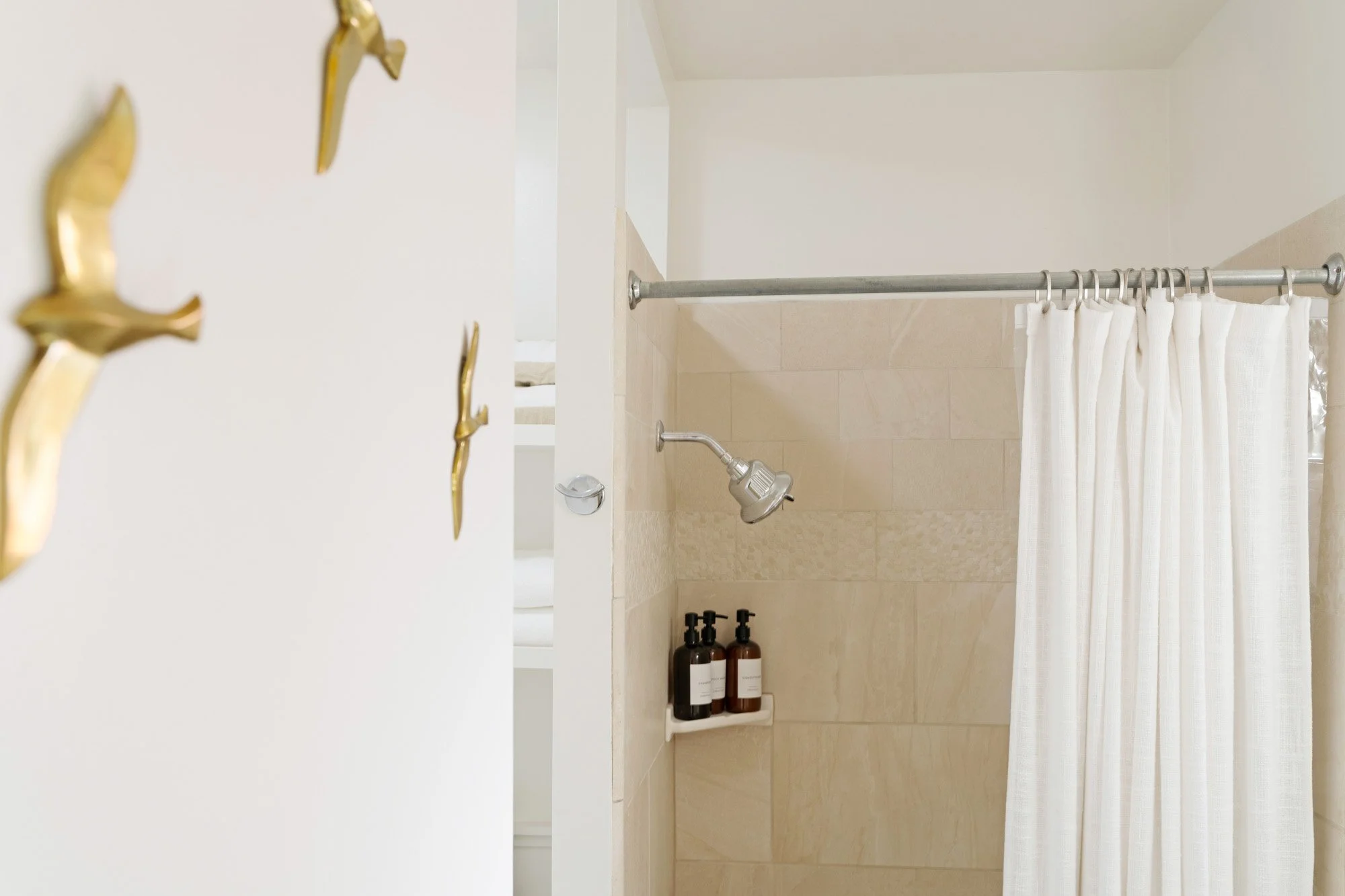 Part of a bathroom with a shower area, featuring a beige tiled wall, a showerhead, a small shelf with bottles, a white curtain, and a white wall with gold hooks.