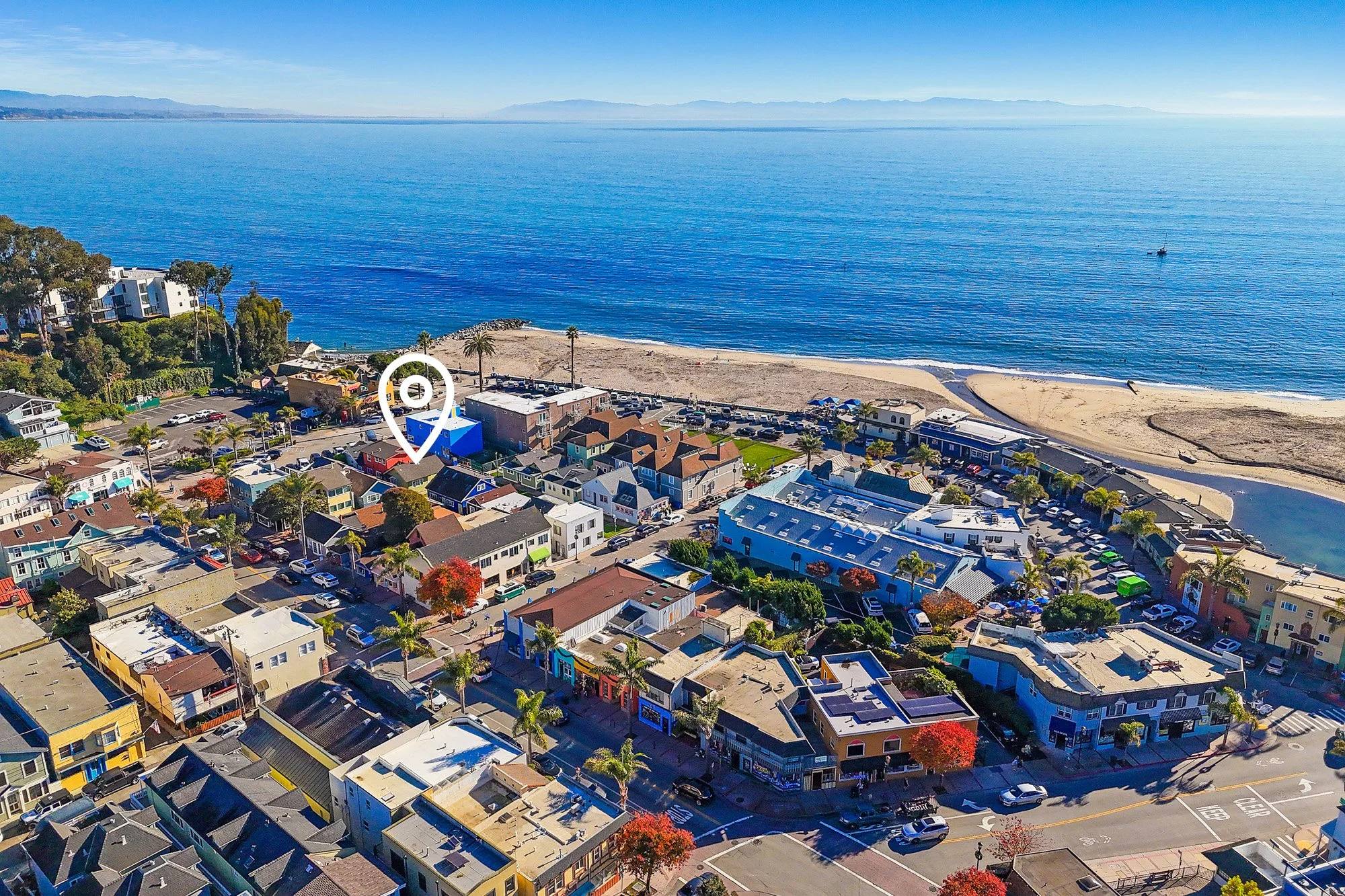 Aerial view of a coastal town with colorful buildings, a beach, and the ocean. The ocean is calm, with a small boat visible. The beach has a paved walkway and a few people. The town features palm trees and a mix of residential and commercial structur