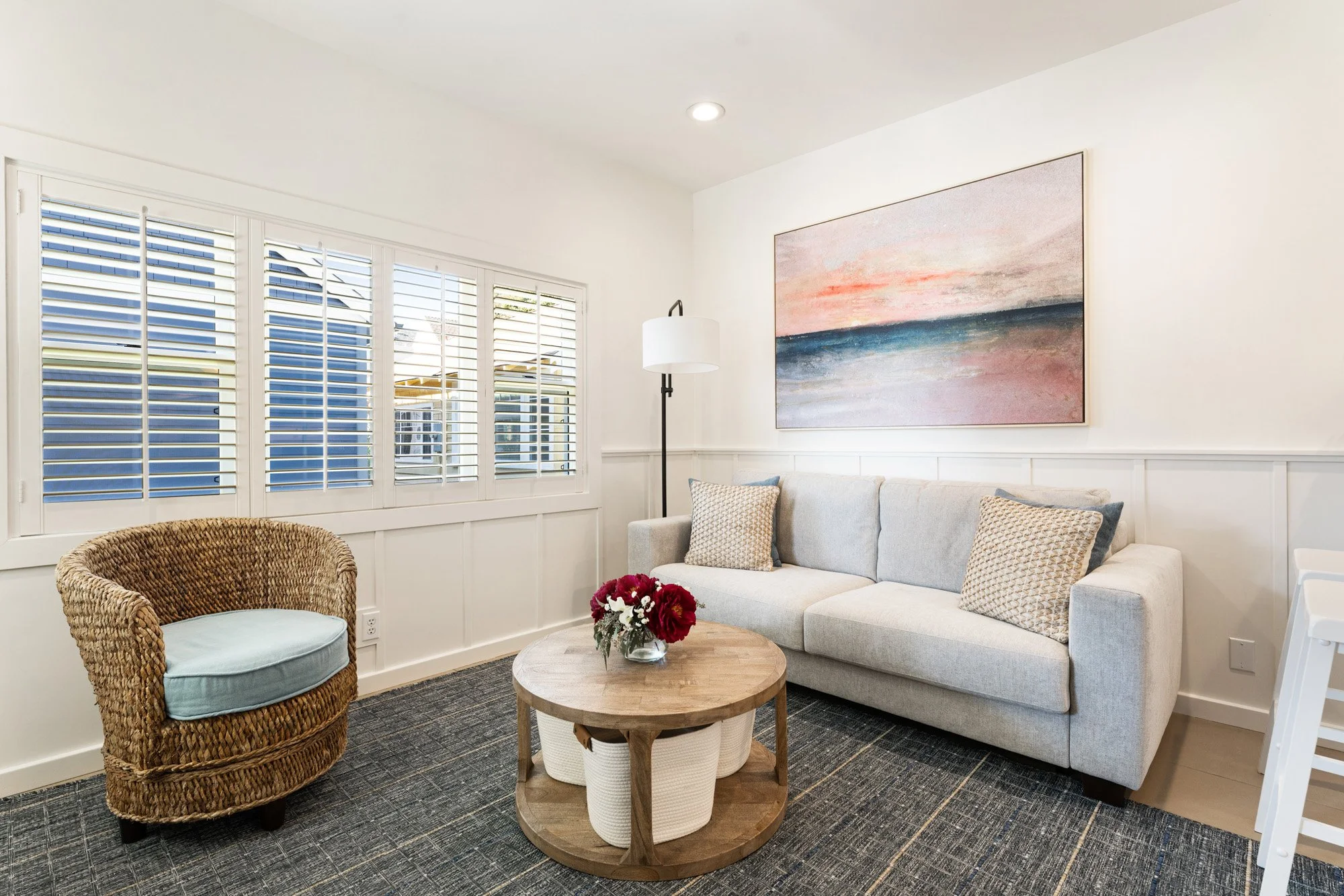 Bright living room with white walls, a large window with white shutters, a beige sofa with patterned pillows, a wicker armchair with a cushion, a round wooden coffee table with flower arrangement, a tall floor lamp, and a colorful abstract painting h