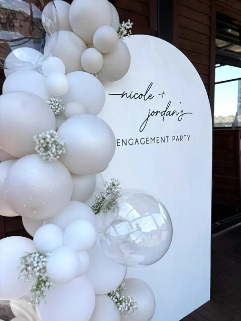 White balloons clustered around a white vertical sign with text for Nicole and Jordan's engagement party.
