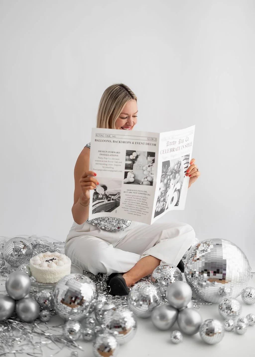 A woman sitting on the floor surrounded by silver disco balls, reading a newspaper, with a birthday cake nearby, in a festive setting.