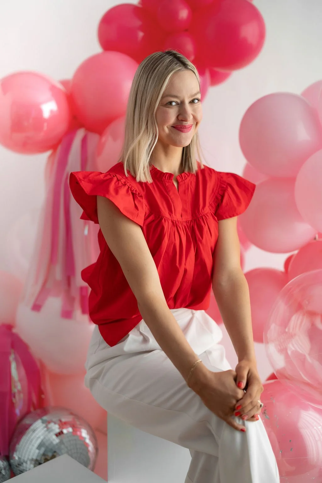 A woman with shoulder-length blonde hair, wearing a red ruffled blouse and white pants, sitting on a white block, smiling with pink balloons and pink streamers in the background.