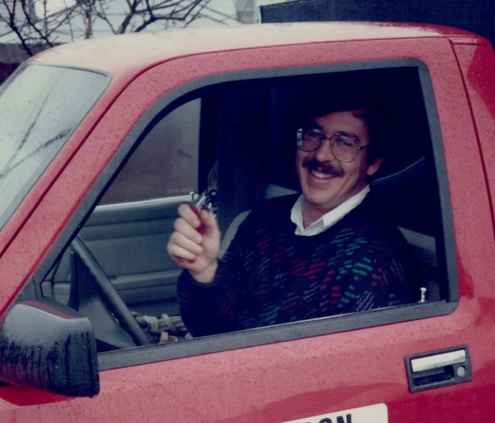 A man with glasses and a mustache smiling and giving a thumbs-up from the window of a red vehicle.