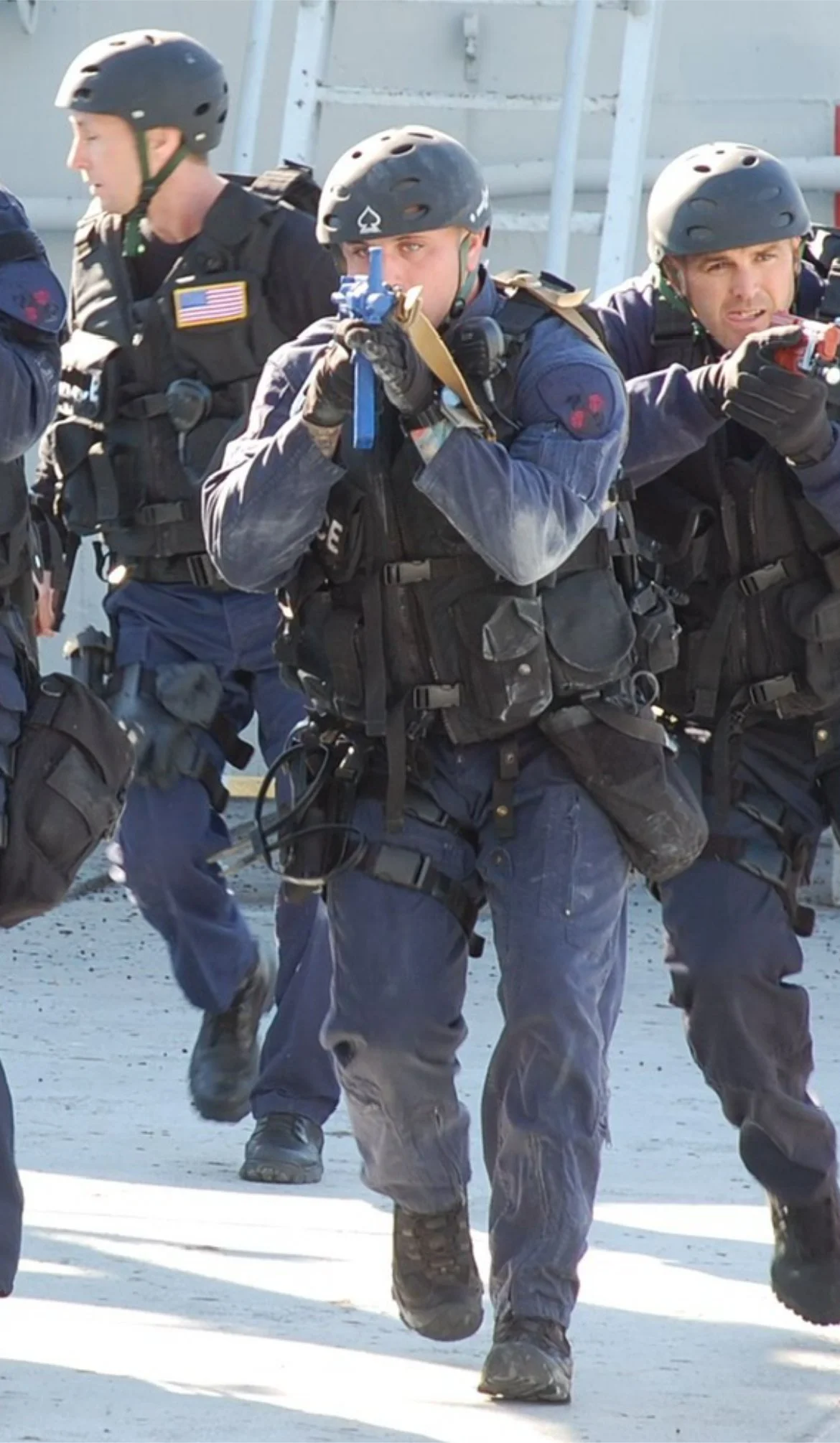 A group of police officers in tactical gear, wearing helmets, and black uniforms, running outdoors with a serious expression.