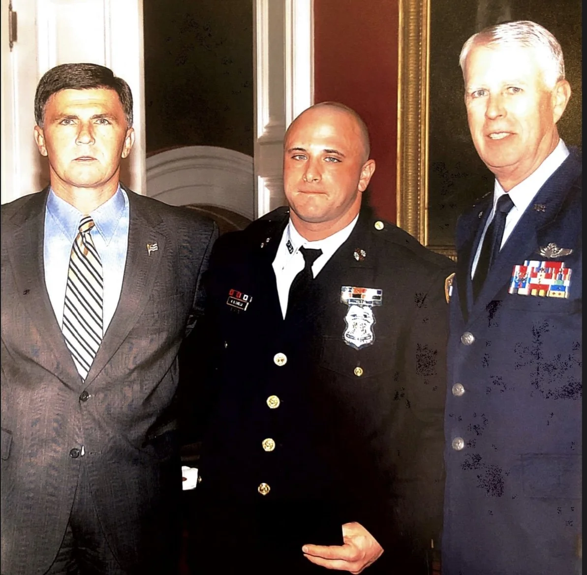 Three men in formal attire and military uniform posing for a photo indoors. The man on the left is in a suit, the man in the middle is in a police uniform, and the man on the right is in a military uniform with medals.