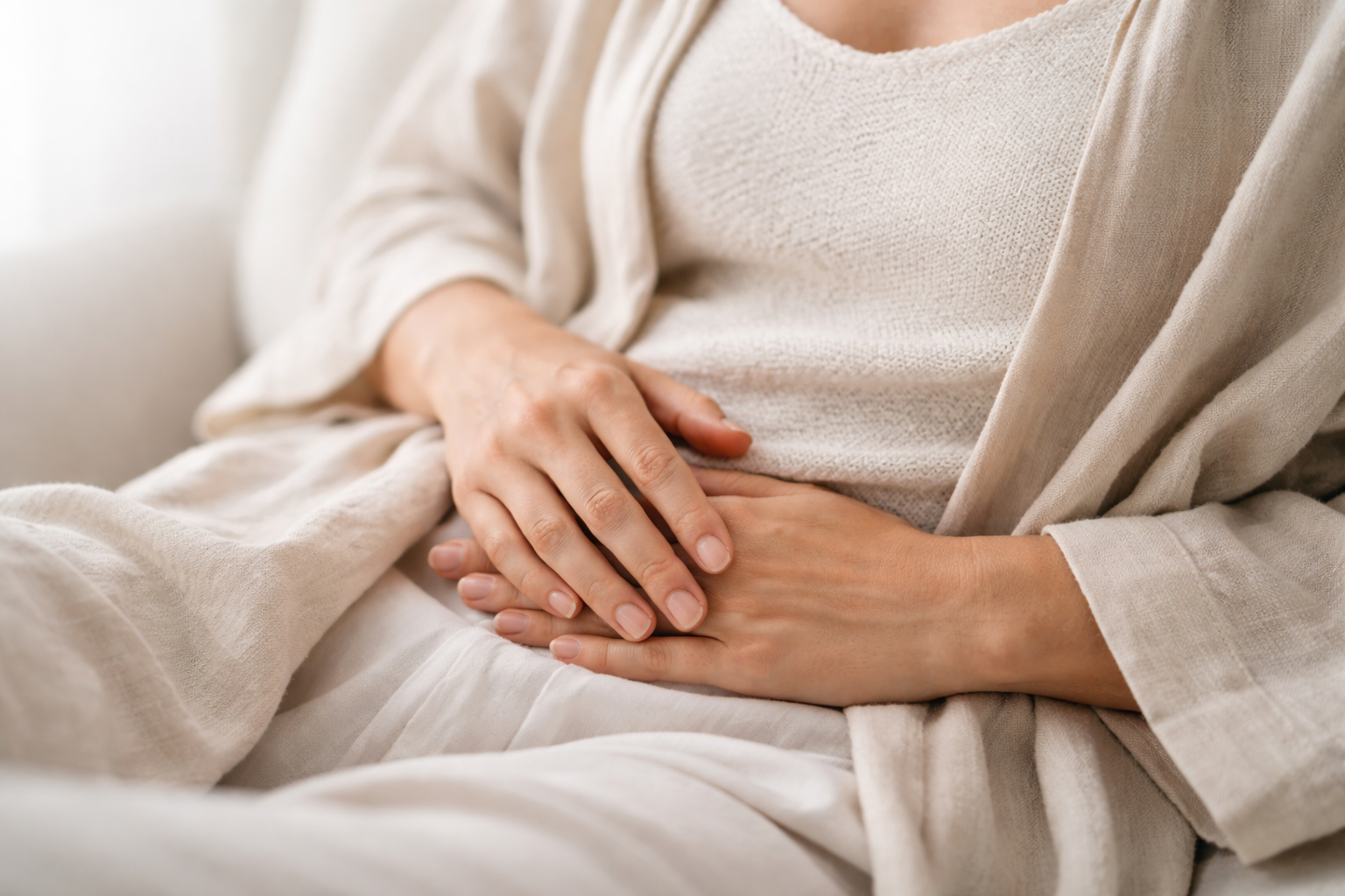Close-up of a person's hands resting on their stomach, wearing light-colored, comfortable clothing, with soft lighting.