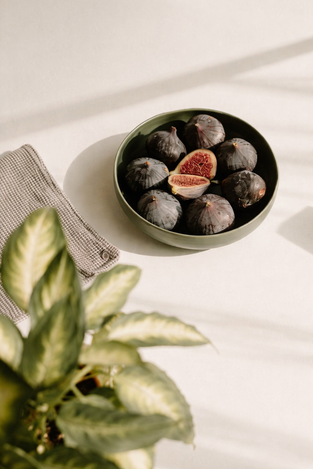 A green bowl filled with fresh black figs, some halved to reveal red flesh, on a white table with a beige woven cloth and a potted plant in the foreground.