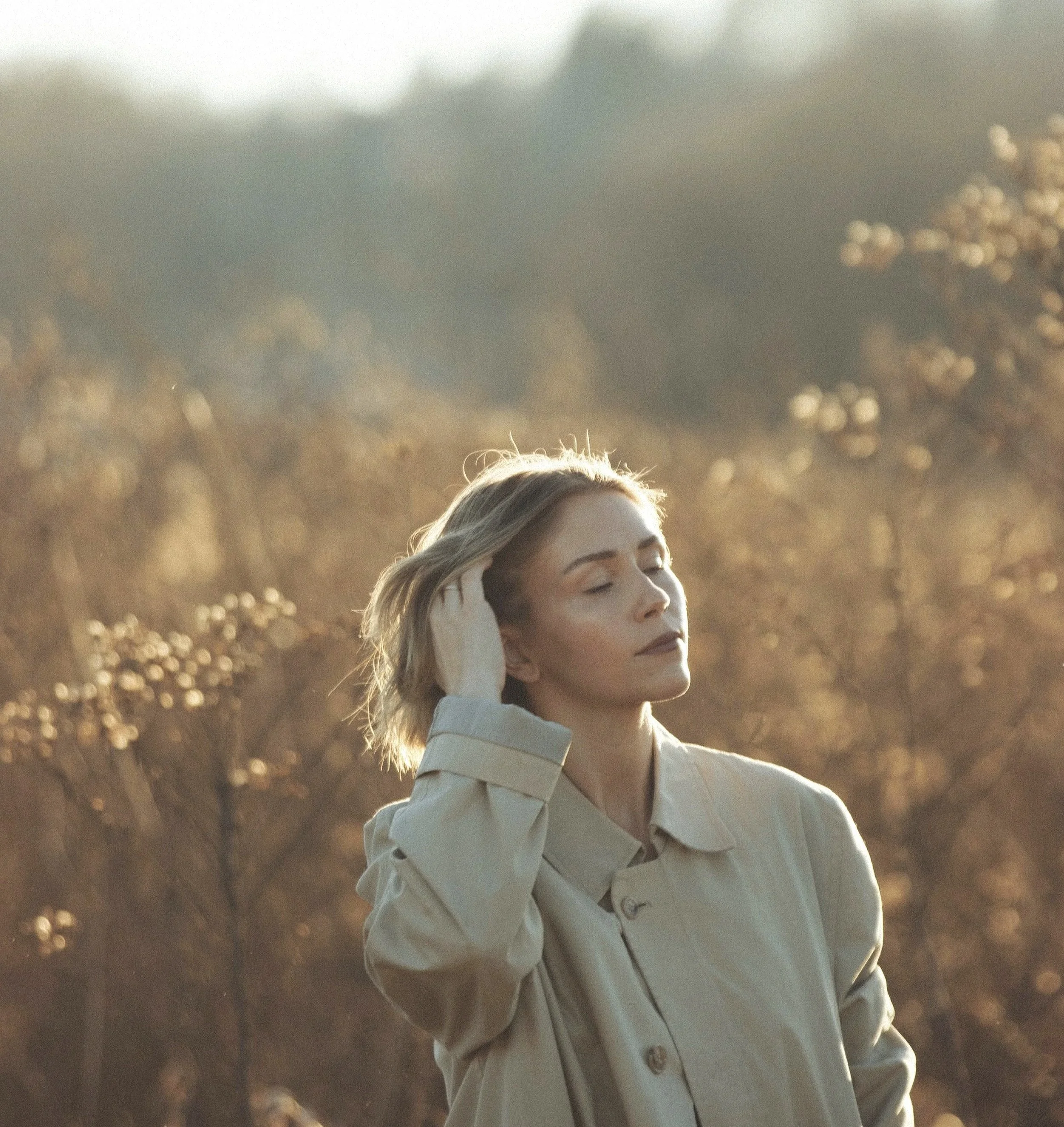 A woman with blonde hair and closed eyes, wearing a beige coat, stands outdoors in a field of trees during sunset.