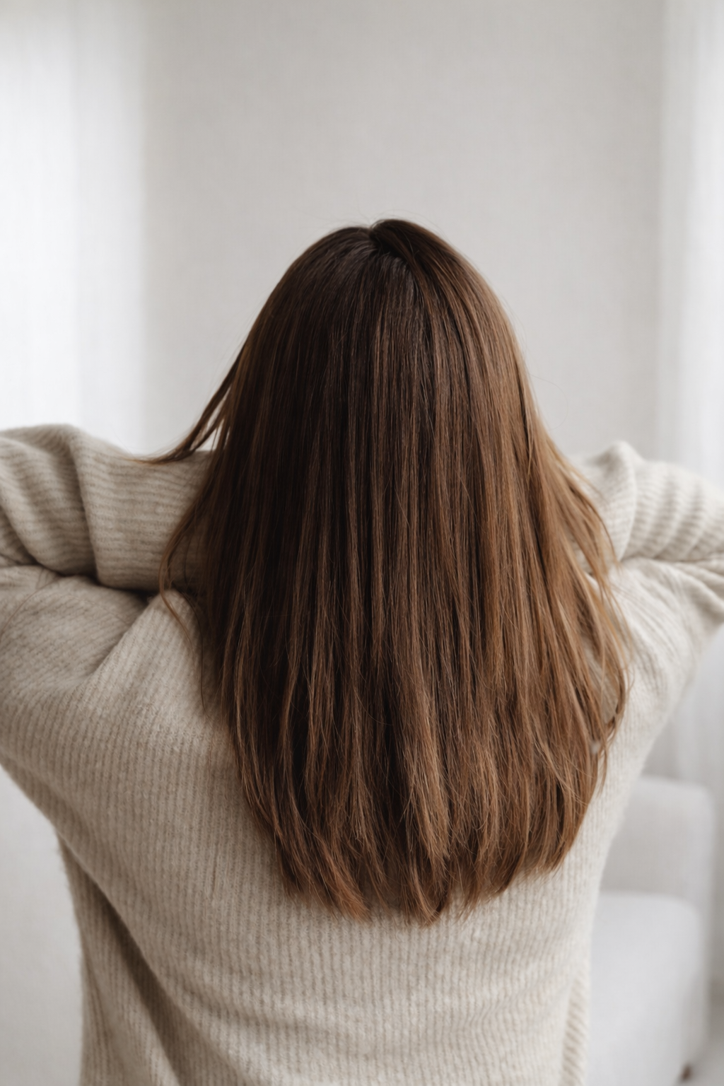 Back of a woman with long, brown hair, wearing a beige knit sweater, stretching her arms with her head down.
