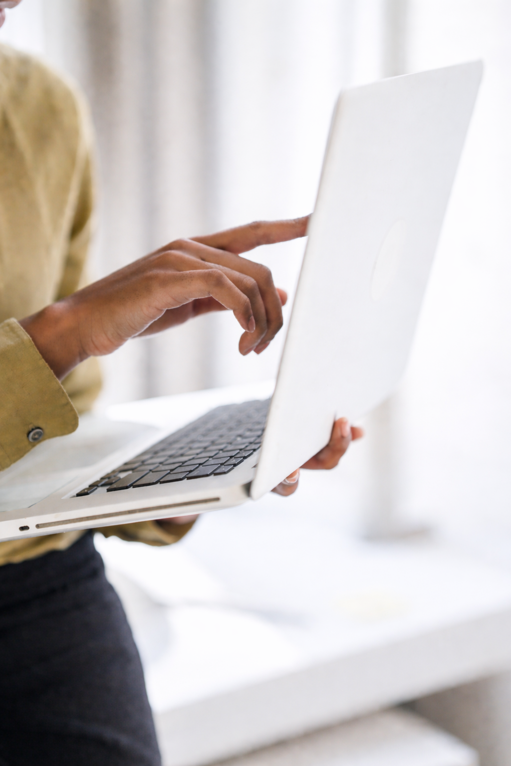 Person holding a white laptop, typing with one hand, in a bright room.