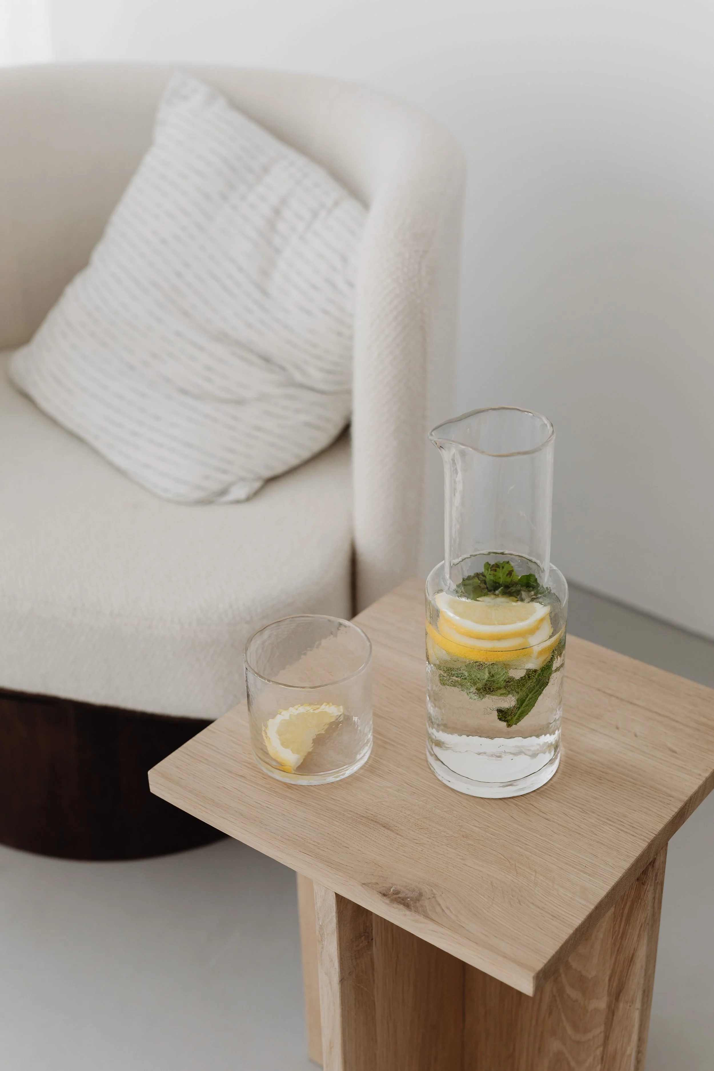 Glass pitcher and small glass cup with lemon slices and mint leaves on a wooden table in a room with beige armchair and striped pillow.