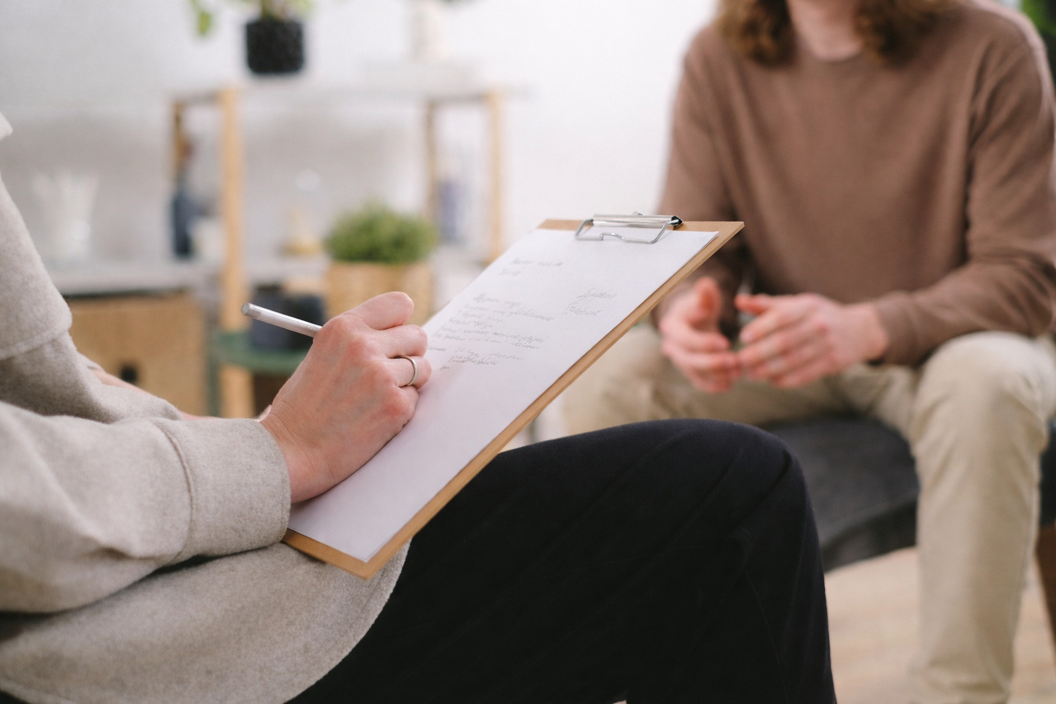 Person taking notes on a clipboard during a medical intake session with a woman sitting on a couch in a bright room.