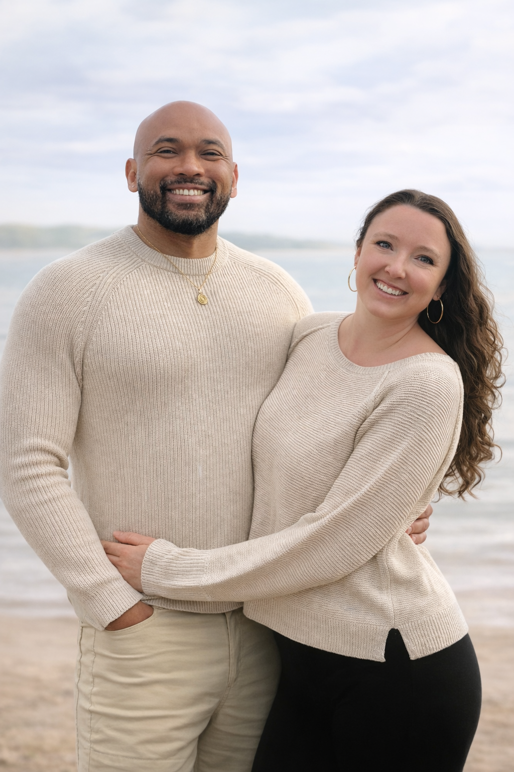 A couple standing on a beach, smiling with arms around each other, with water and cloudy sky in the background.