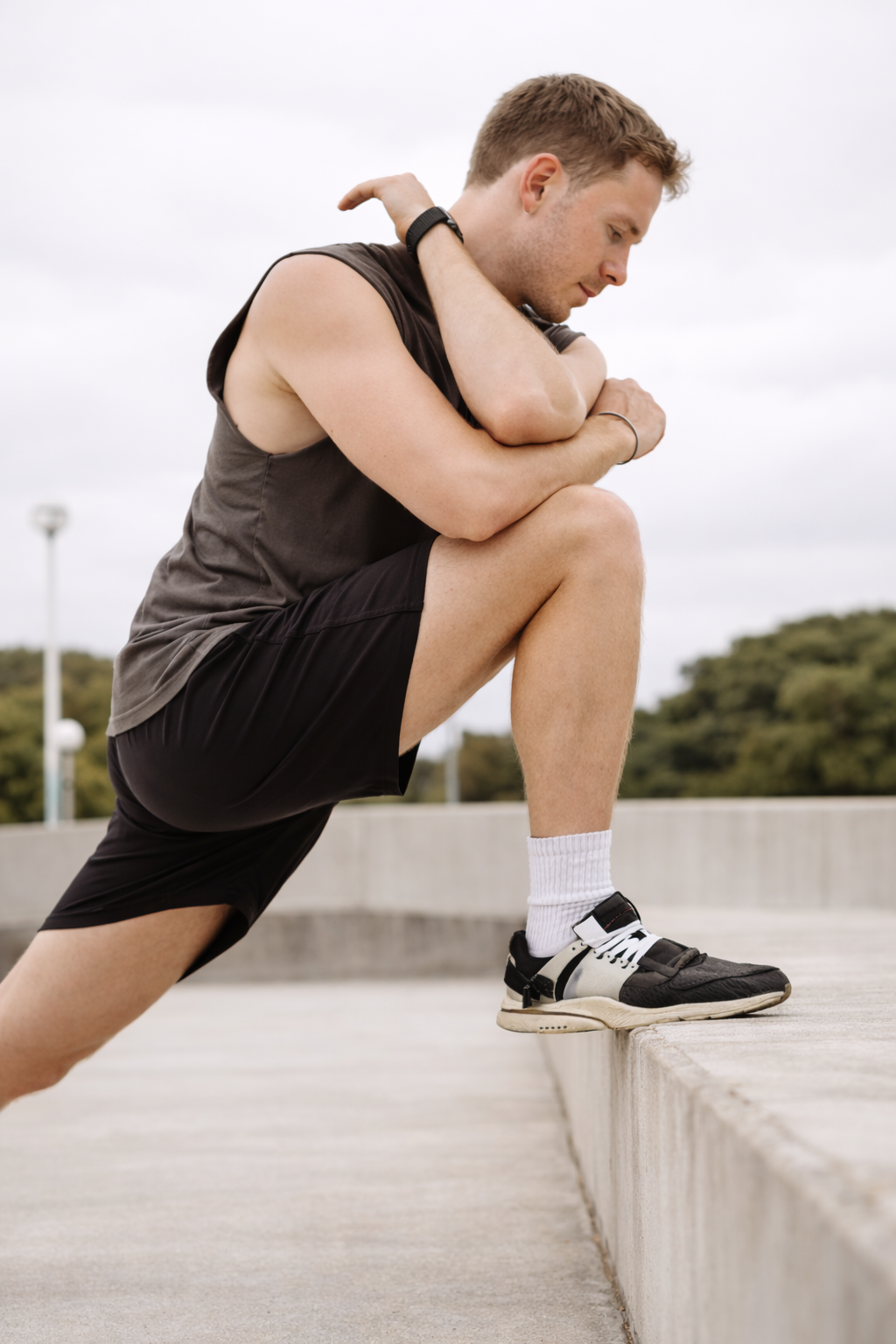 A young man in athletic clothing stretching outdoors, lunging with one leg on a curb and the other extended behind him, with his arms crossed over his knee and looking down, overcast sky in background.
