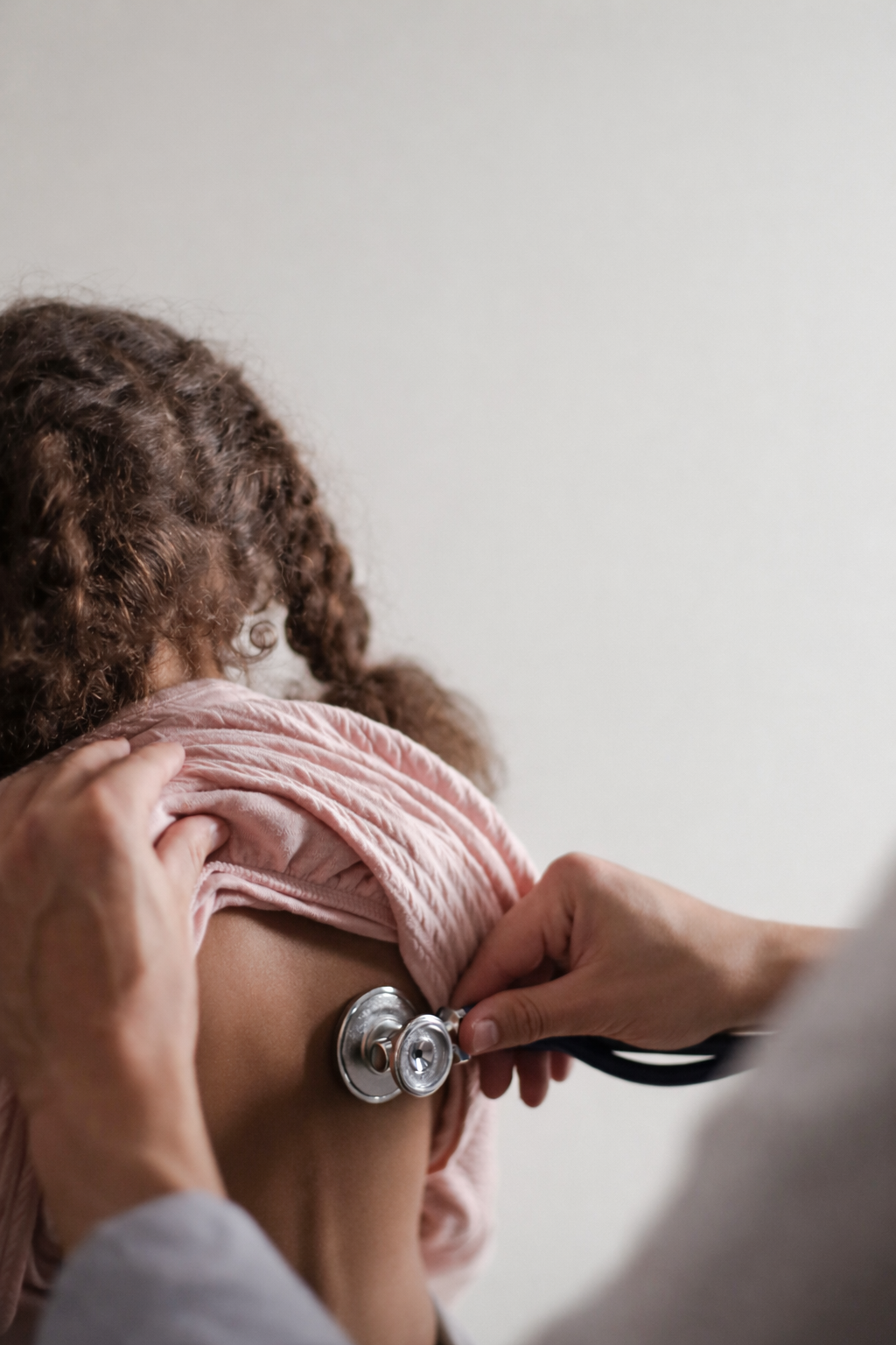 A healthcare professional uses a stethoscope to listen to a young girl's back. The girl wears a pink quilted jacket and has curly brown hair.