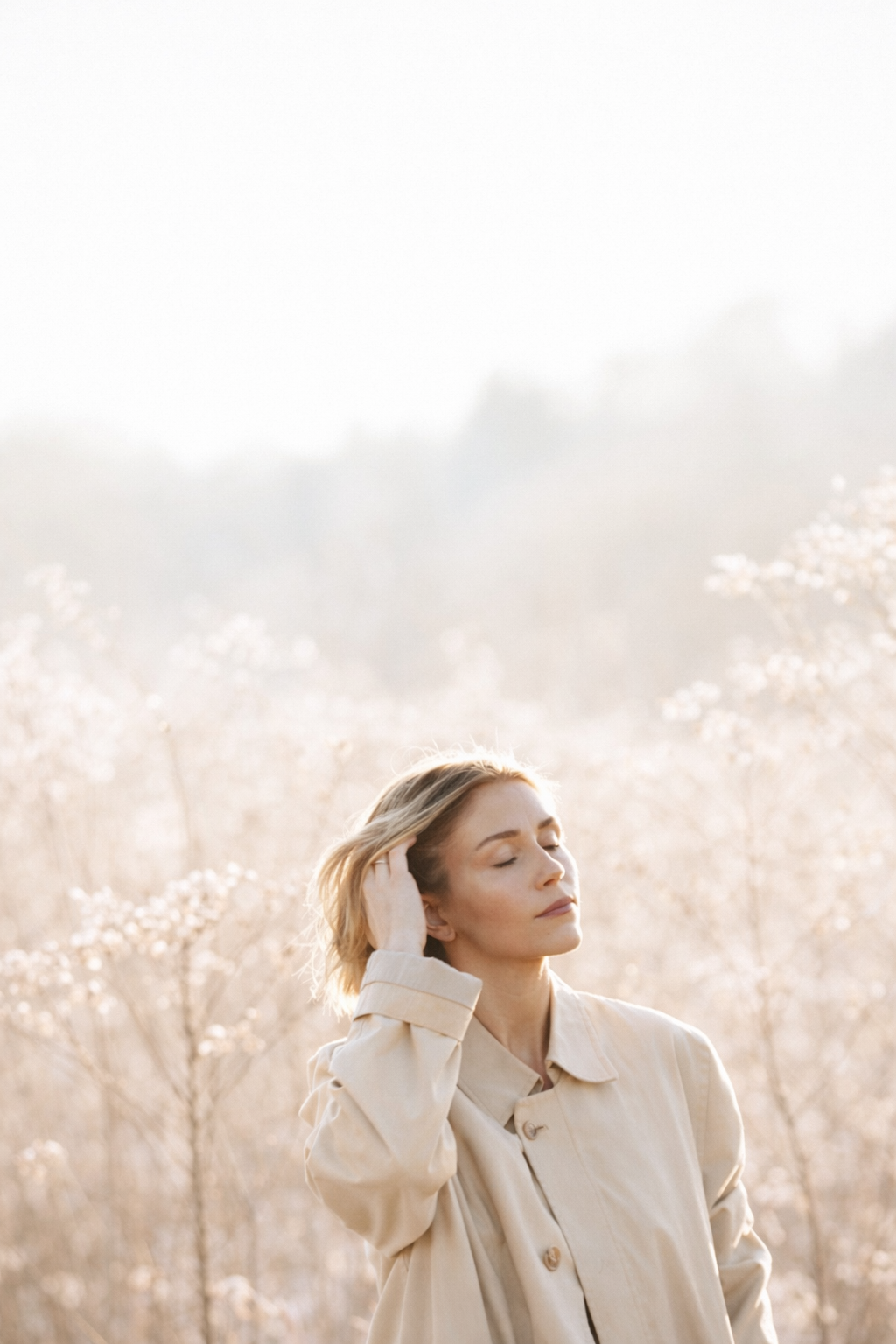 A woman with short blonde hair standing in a field of dry, white flowers, closing her eyes and touching her hair, with soft sunlight illuminating her face and background.