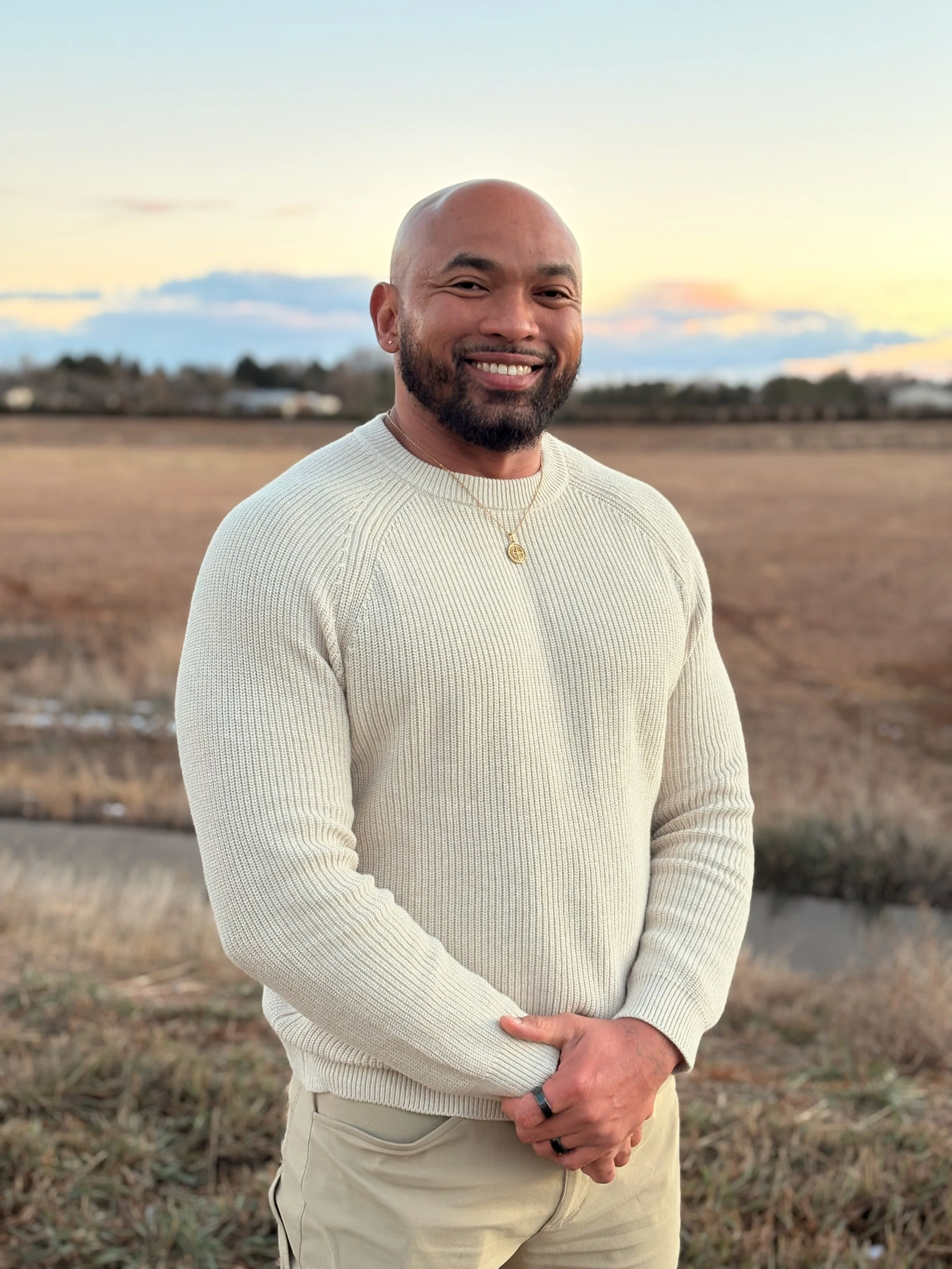 A smiling man with a beard and a gold necklace standing outdoors in a field during sunset.